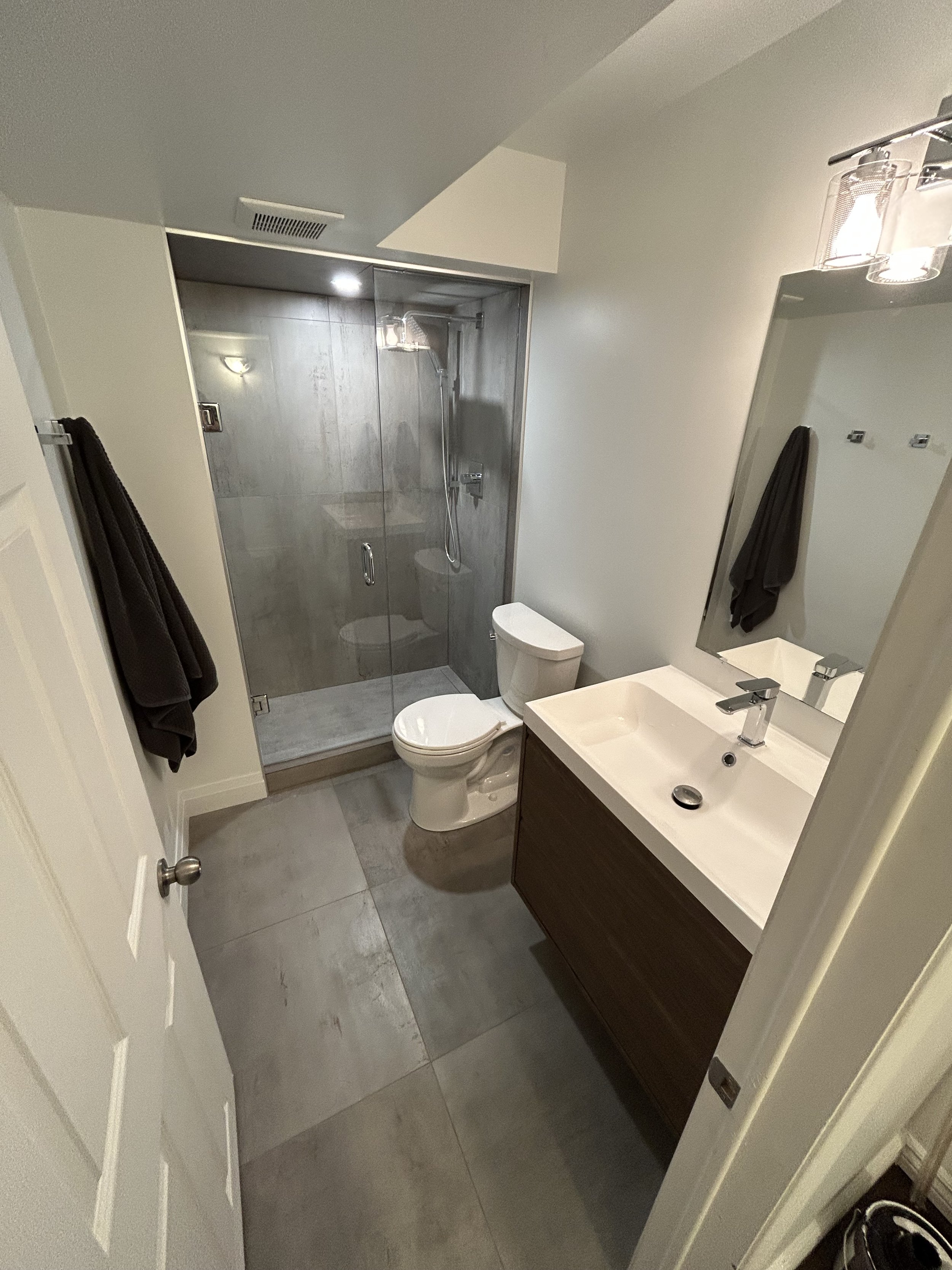Bathroom with a shower, toilet, sink with mirror, dark towel, and gray tile floor.