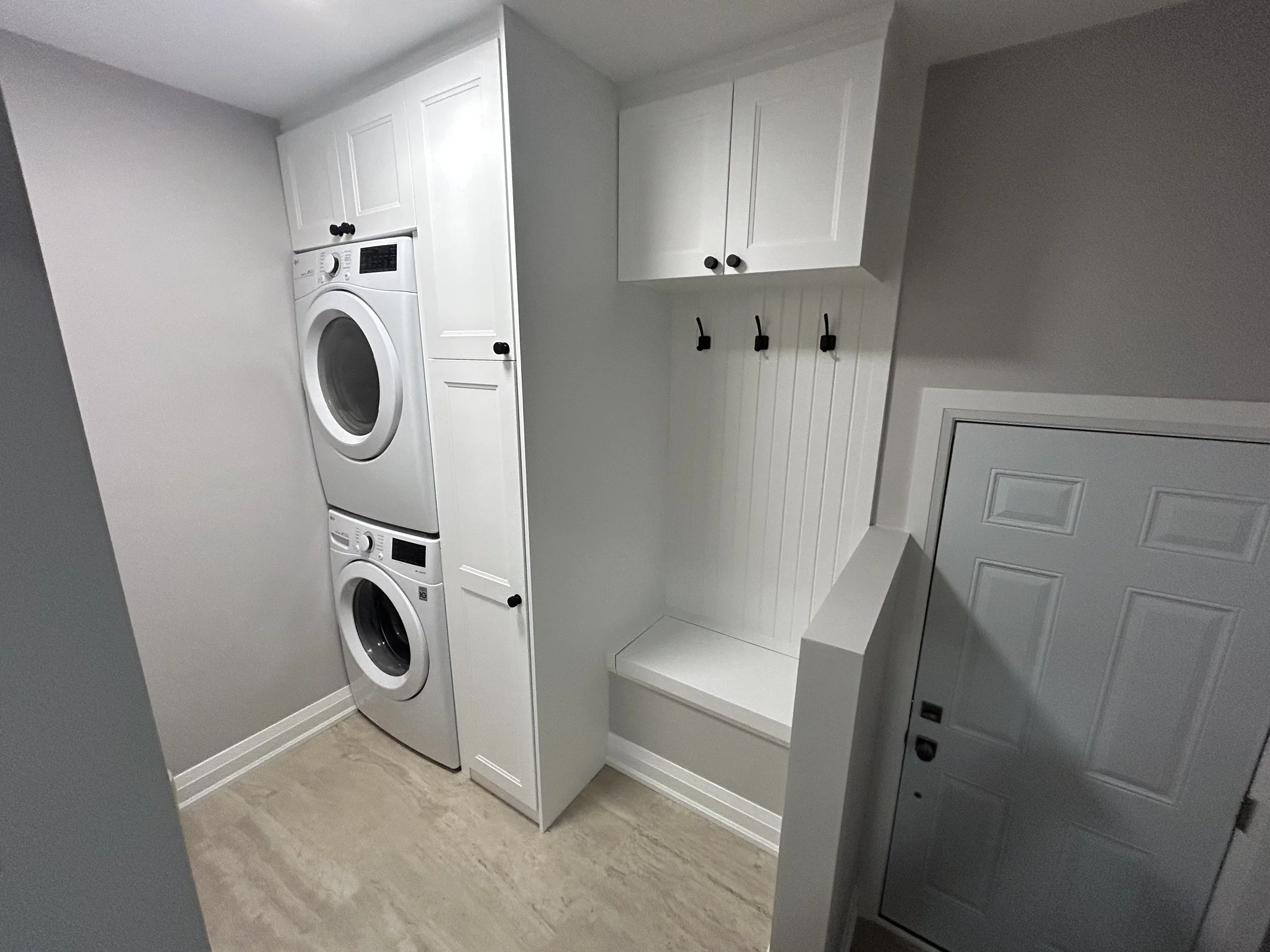A laundry room with a front-loading washing machine and dryer stacked vertically, white cabinetry above, and a white wainscot panel on the wall. The room has light-colored flooring and neutral-colored walls.
