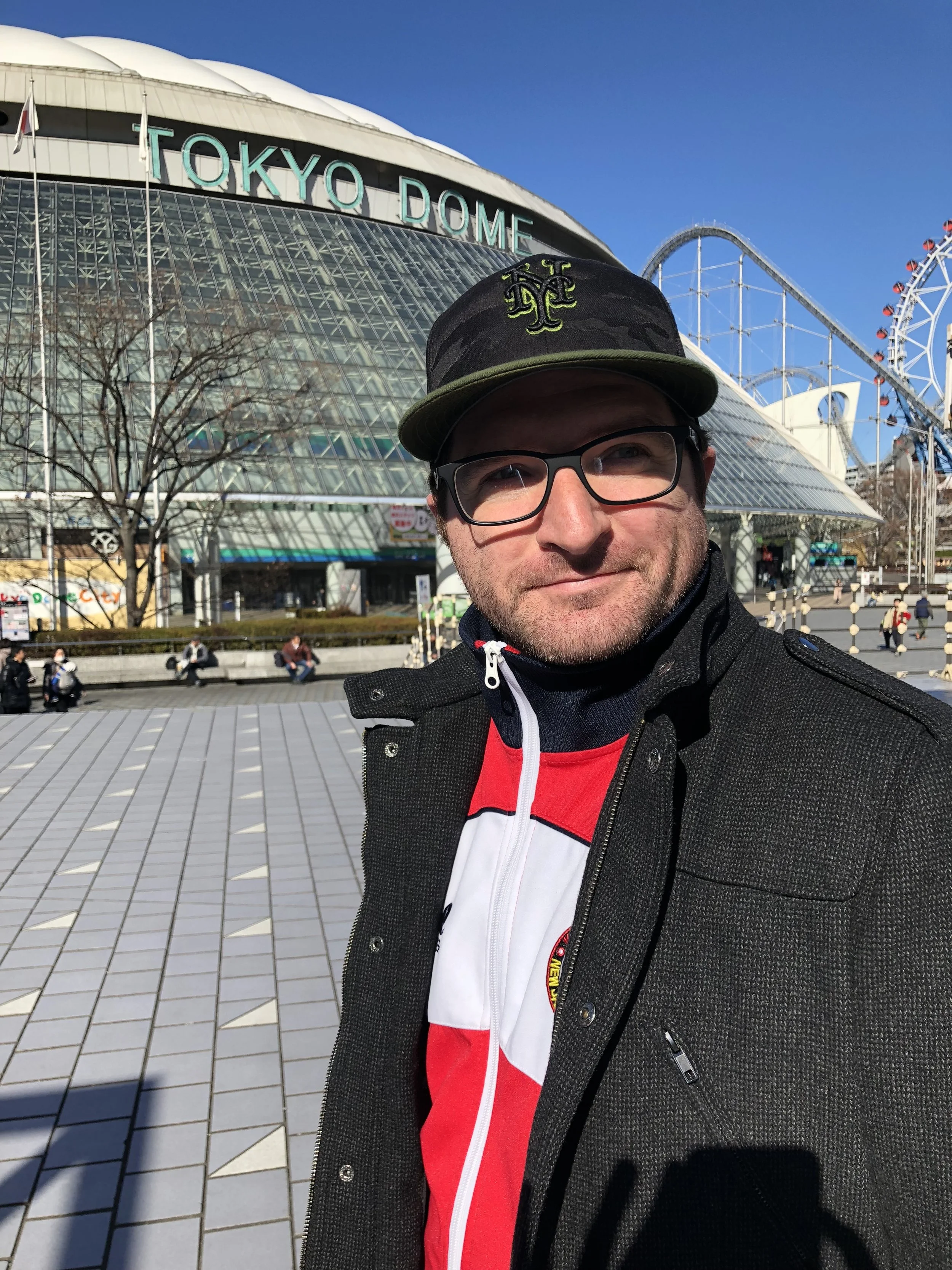 A man taking a selfie in front of the Tokyo Dome with the iconic structure and amusement park rides in the background. He is wearing glasses, a black coat, a black and green cap, and a red, white, and black jacket.