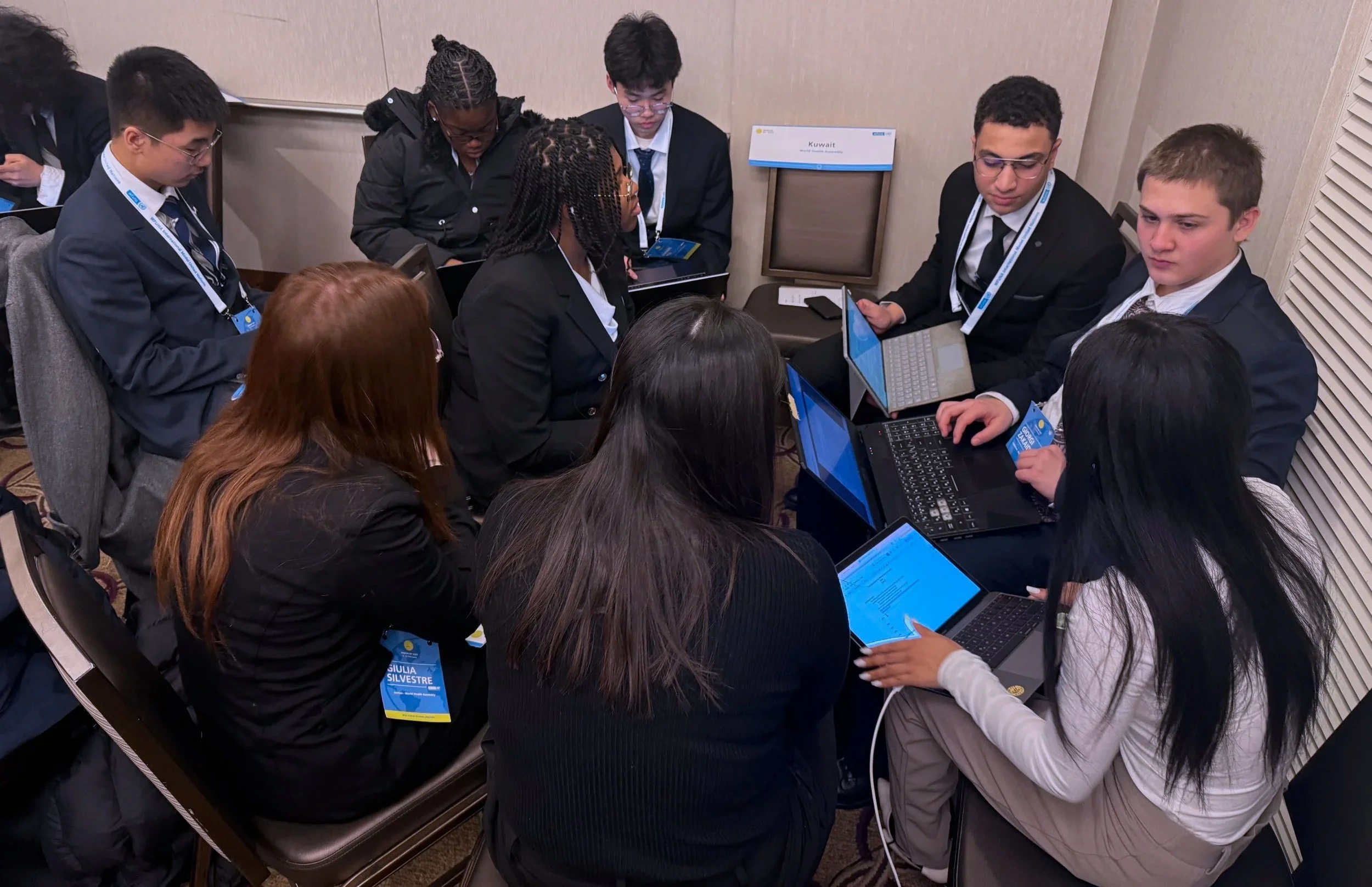 A group of young people in formal attire sitting and standing around a cluster of laptops and tablets during a conference, with name tags hanging around their necks.