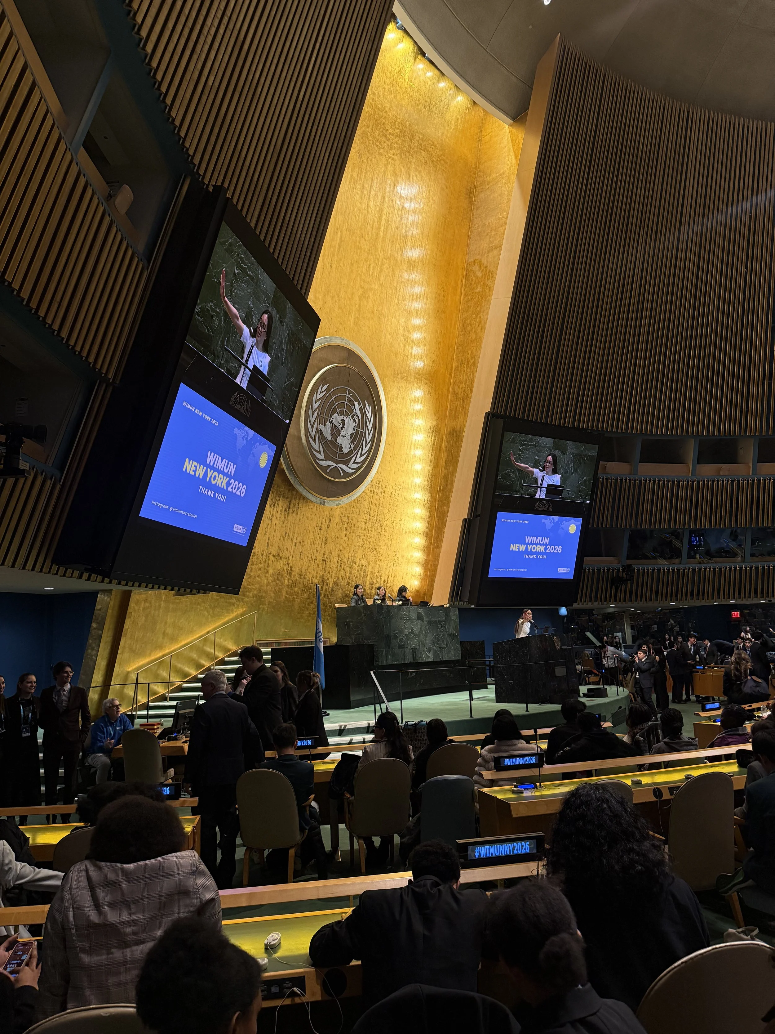 The United Nations Assembly Hall with a speaker at the podium and a large display screen that reads 'WIMUN NEW YORK 2026 THANK YOU!' The hall is filled with attendees sitting at desks, some using laptops or phones, and officials gathered around the stage.