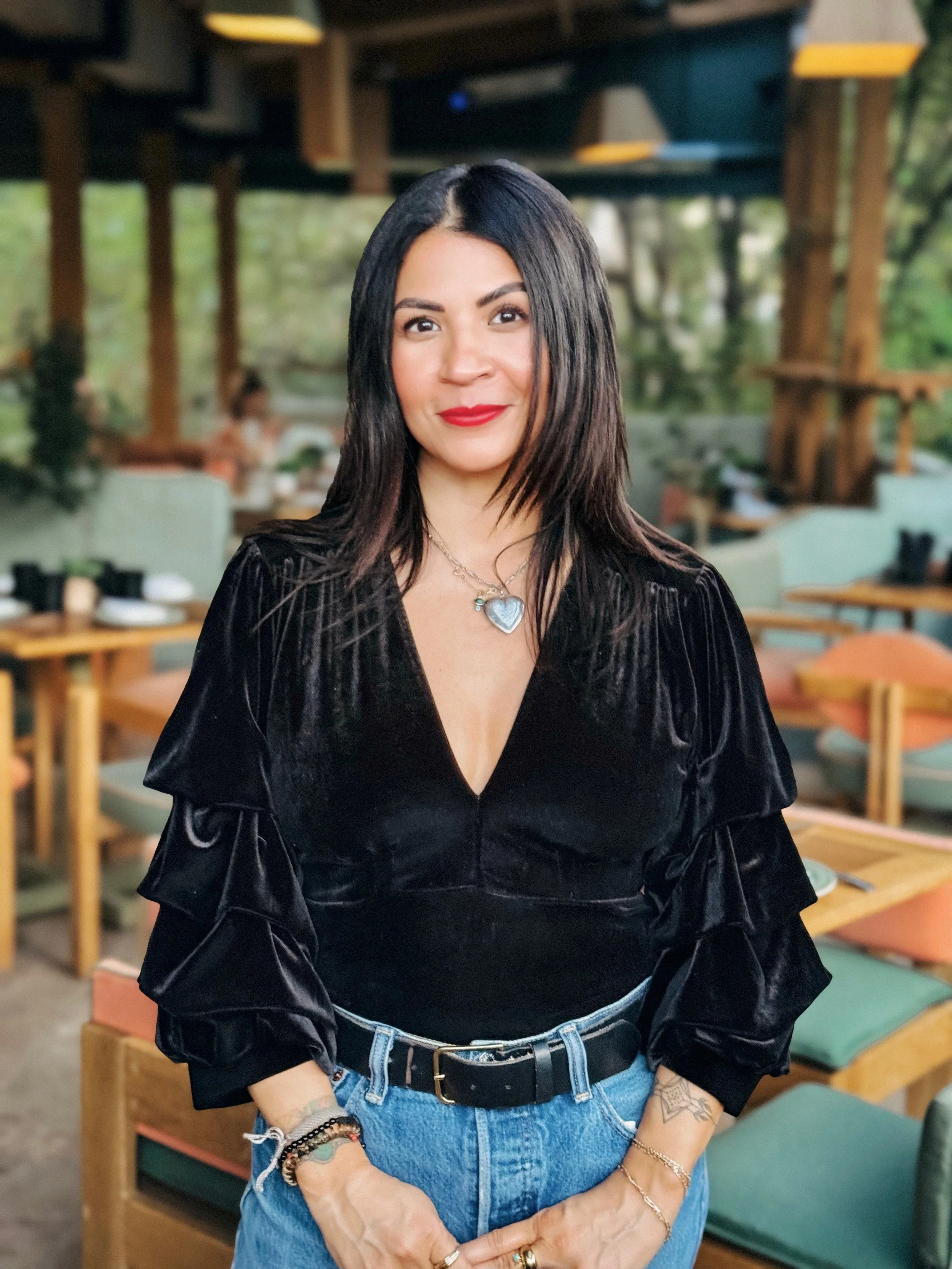A woman with long dark hair and red lipstick standing in a restaurant with wooden tables and green chairs, wearing a black velvet top and blue jeans, accessorized with necklaces and bracelets.