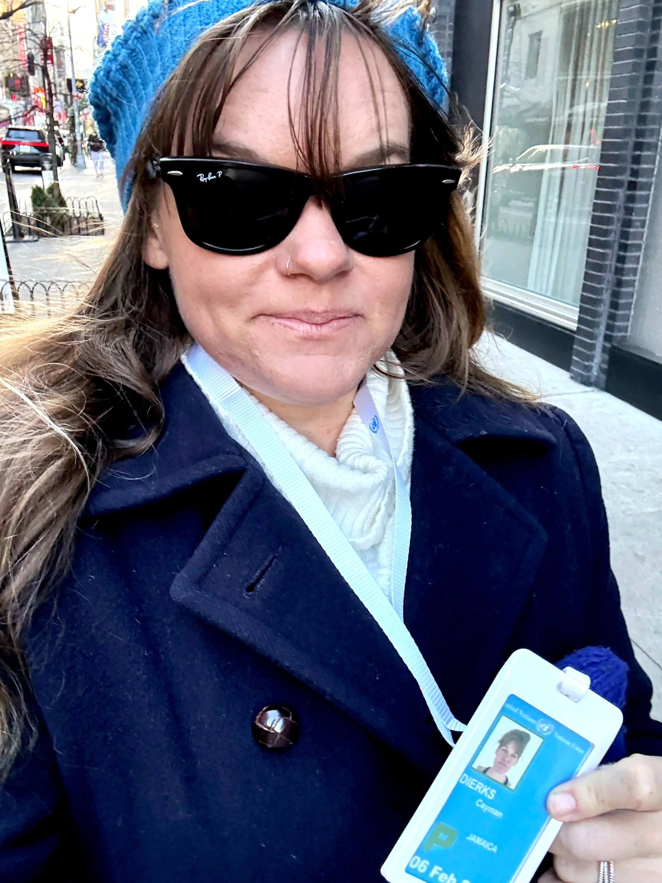 A woman with long brown hair, wearing sunglasses and a blue knit hat, holds up her identification card outside in an urban area with brick buildings and parked cars.