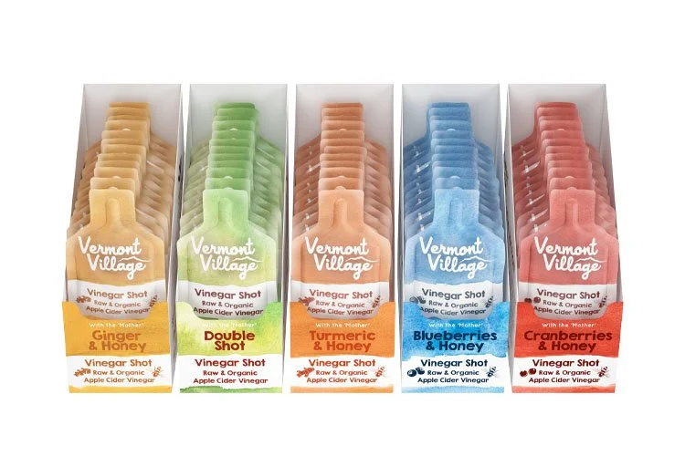 Five boxes of Vermont Village vinegar shot with different flavors and colors, including ginger and honey, double shot, turmeric and honey, blueberries and honey, and cranberries and honey.
