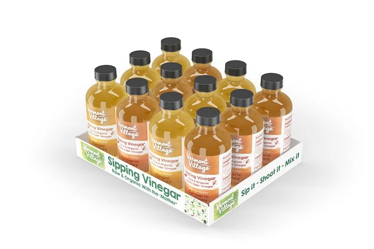 A display box with 15 small bottles of apple cider vinegar, labeled "Vermont Village" and "Sipping Vinegar," arranged in three rows of five, with a tag that says "Sipping Vinegar, low & organic with the Mother, Sip it, Shoot it, Mix it."