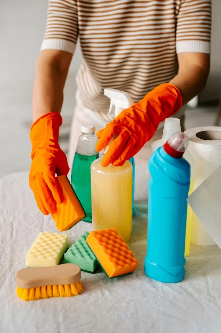 Person wearing orange gloves cleaning various cleaning supplies, including spray bottles, sponges, and a scrub brush, on a white surface.