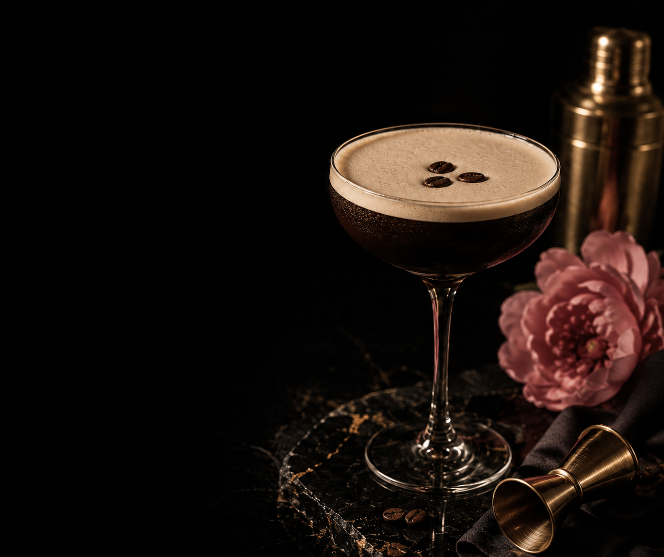 A black cocktail in a coupe glass garnished with coffee beans, placed on a dark marble surface with pink flowers, a gold cocktail shaker, and a gold jigger against a black background.