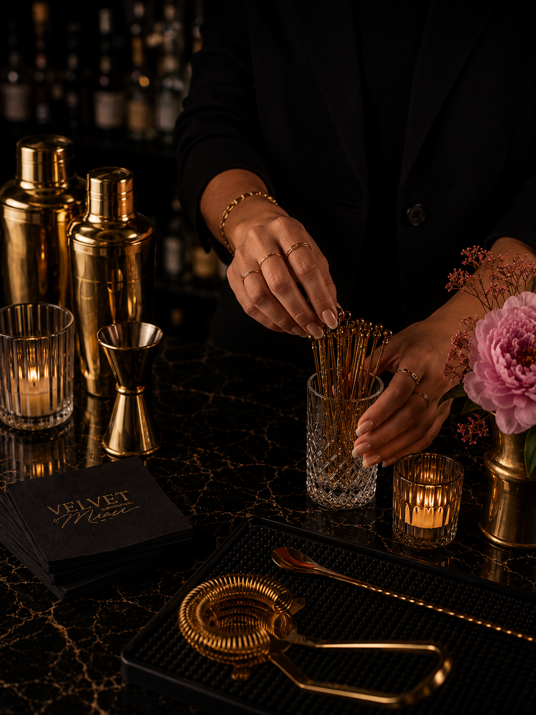 A person in black clothing arranging gold cocktail picks in a glass on a dark marble bar counter with gold and glass barware, flowers, and a menu.
