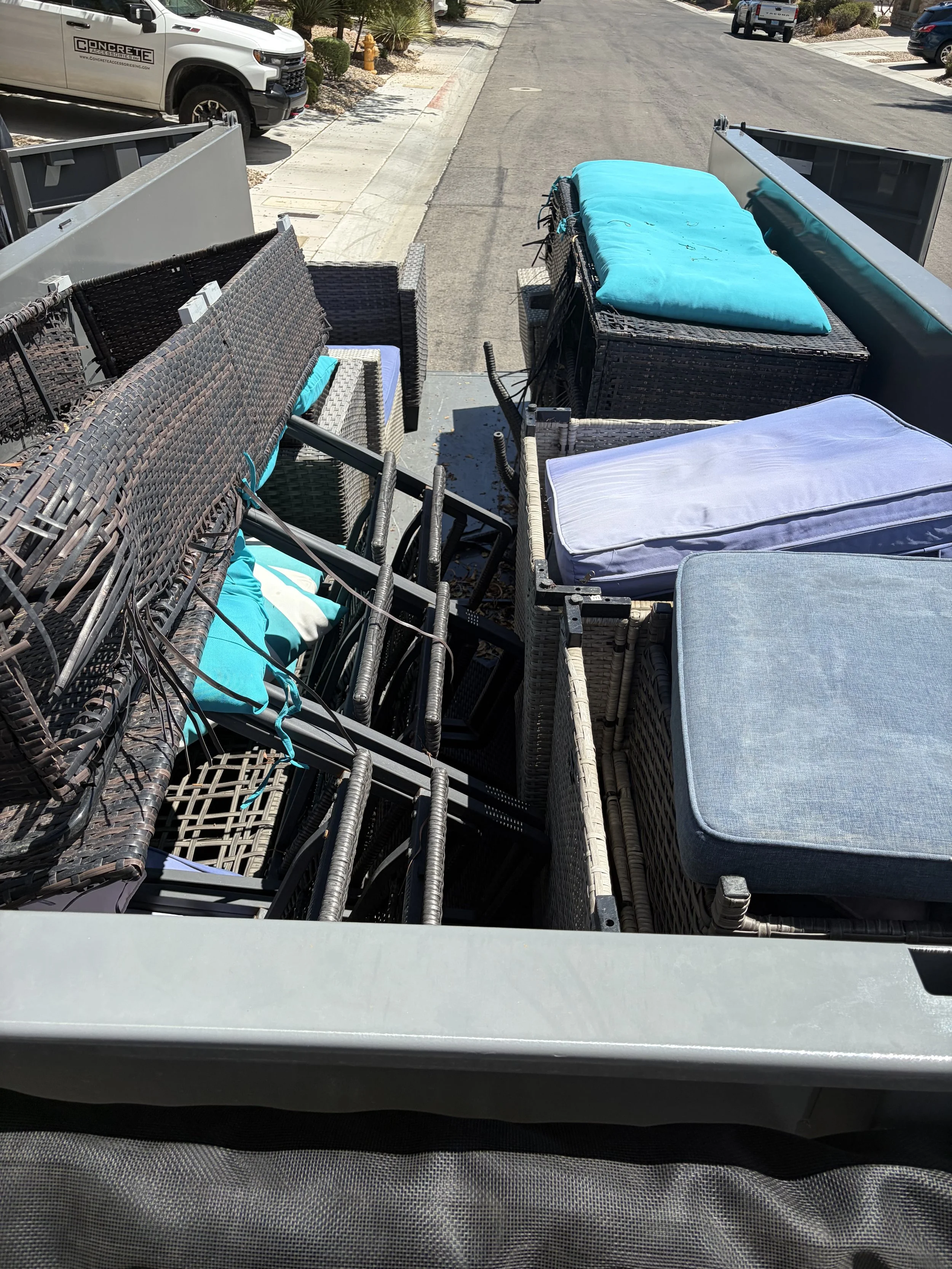 The image shows the back of a truck filled with outdoor furniture, including chairs, cushions, and tables, parked on a residential street.