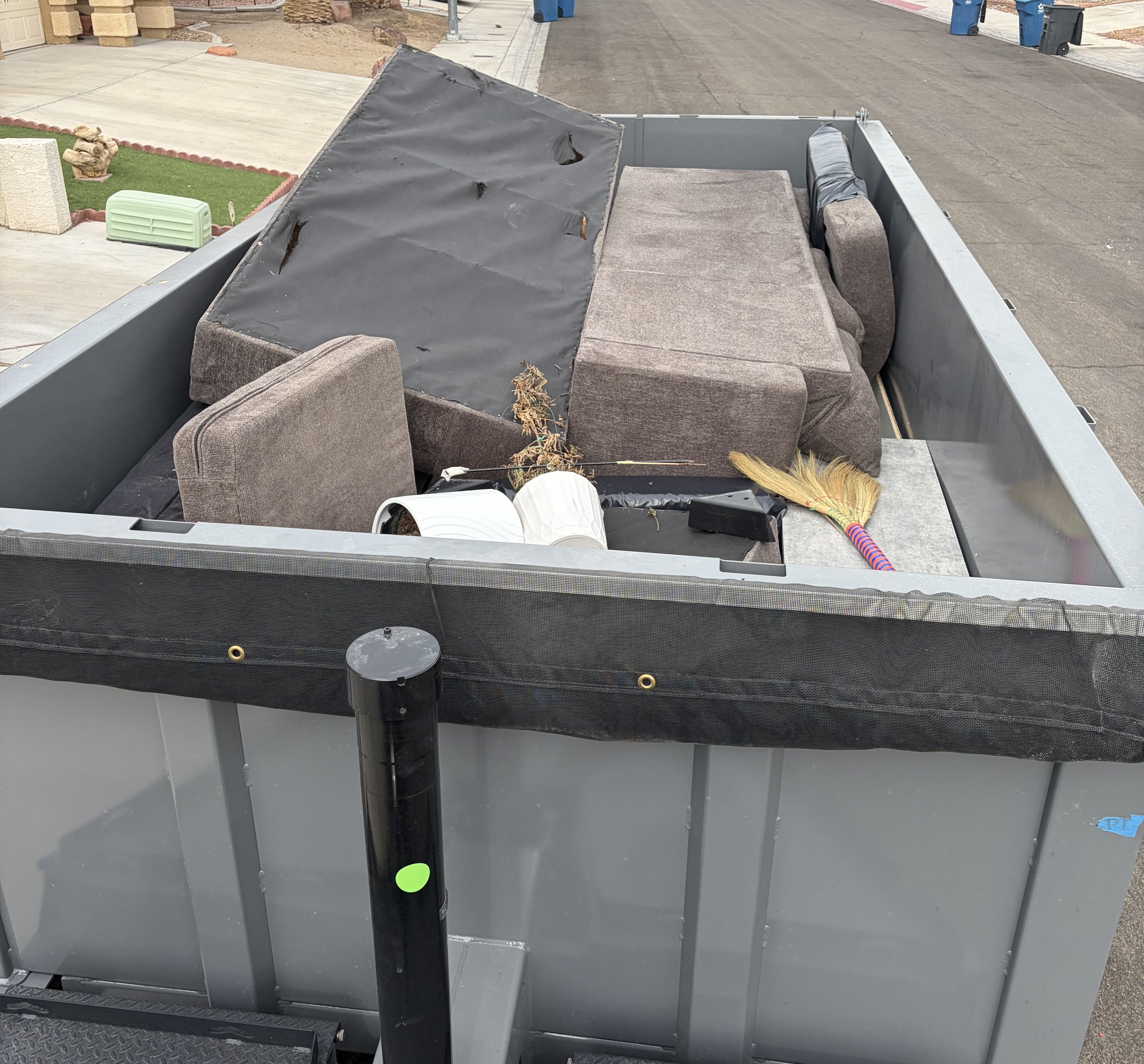 The image shows the truck bed filled with old furniture, including a couch, along with a broom, a white pot, and some miscellaneous items. The scene is set in a residential area with a driveway and street visible in the background.