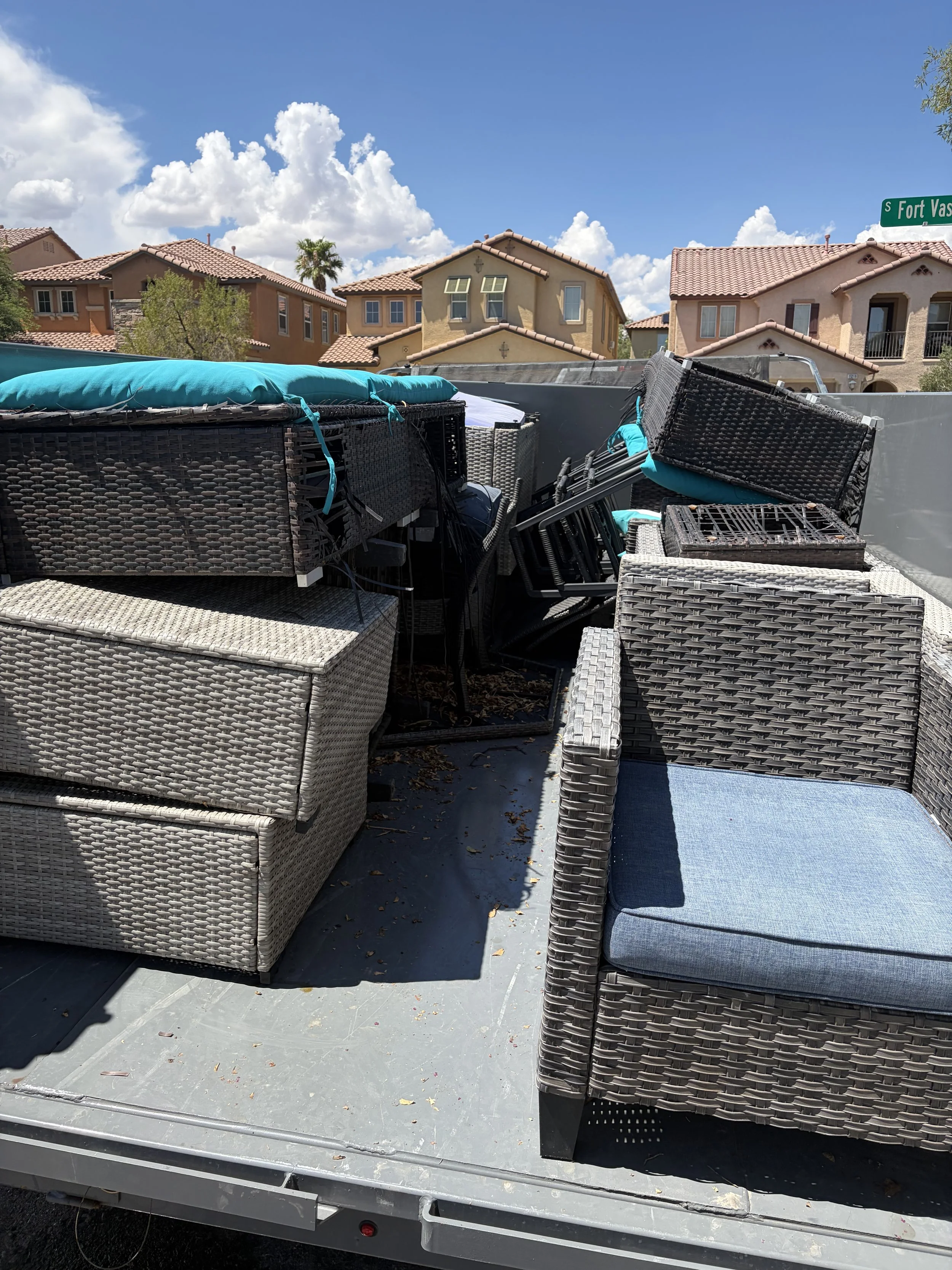 Outdoor scene with stacked patio furniture, including wicker chairs with cushions, on a flatbed trailer under a blue sky with clouds, in a residential neighborhood.