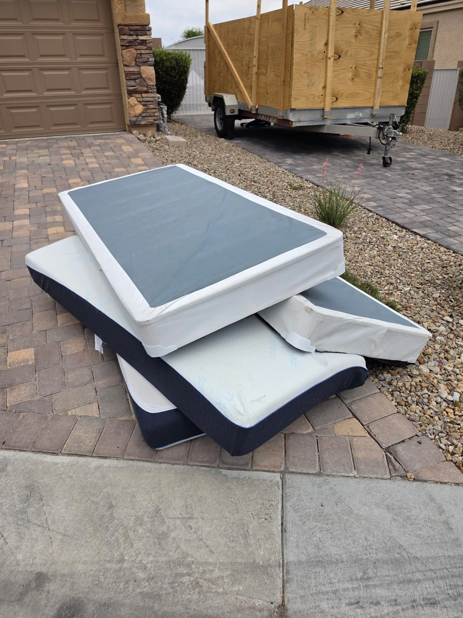 Two mattresses stacked on top of each other on a paved driveway, with some weathered concrete sidewalk steps in the foreground, and a trailer with wood planks in the background.