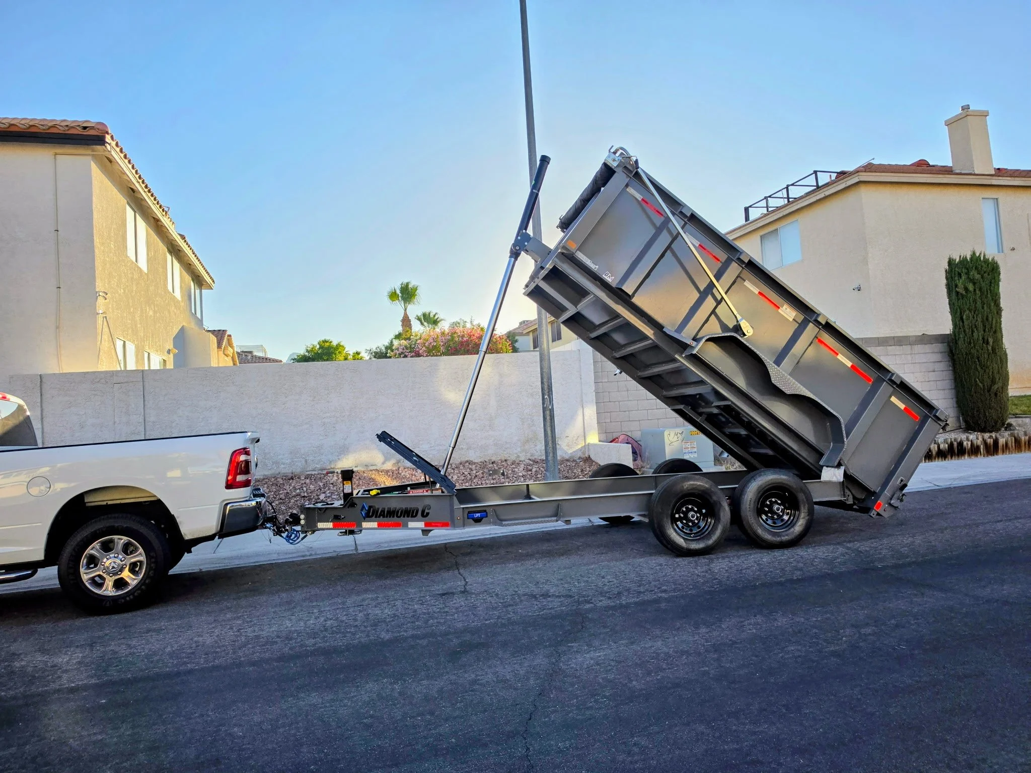 A white pickup truck with a flatbed towing trailer attached, which is raised at an angle, situated on a residential street. In the background, there are two-story houses with beige walls, tiled roofs, and palm trees. The sky is clear and blue.