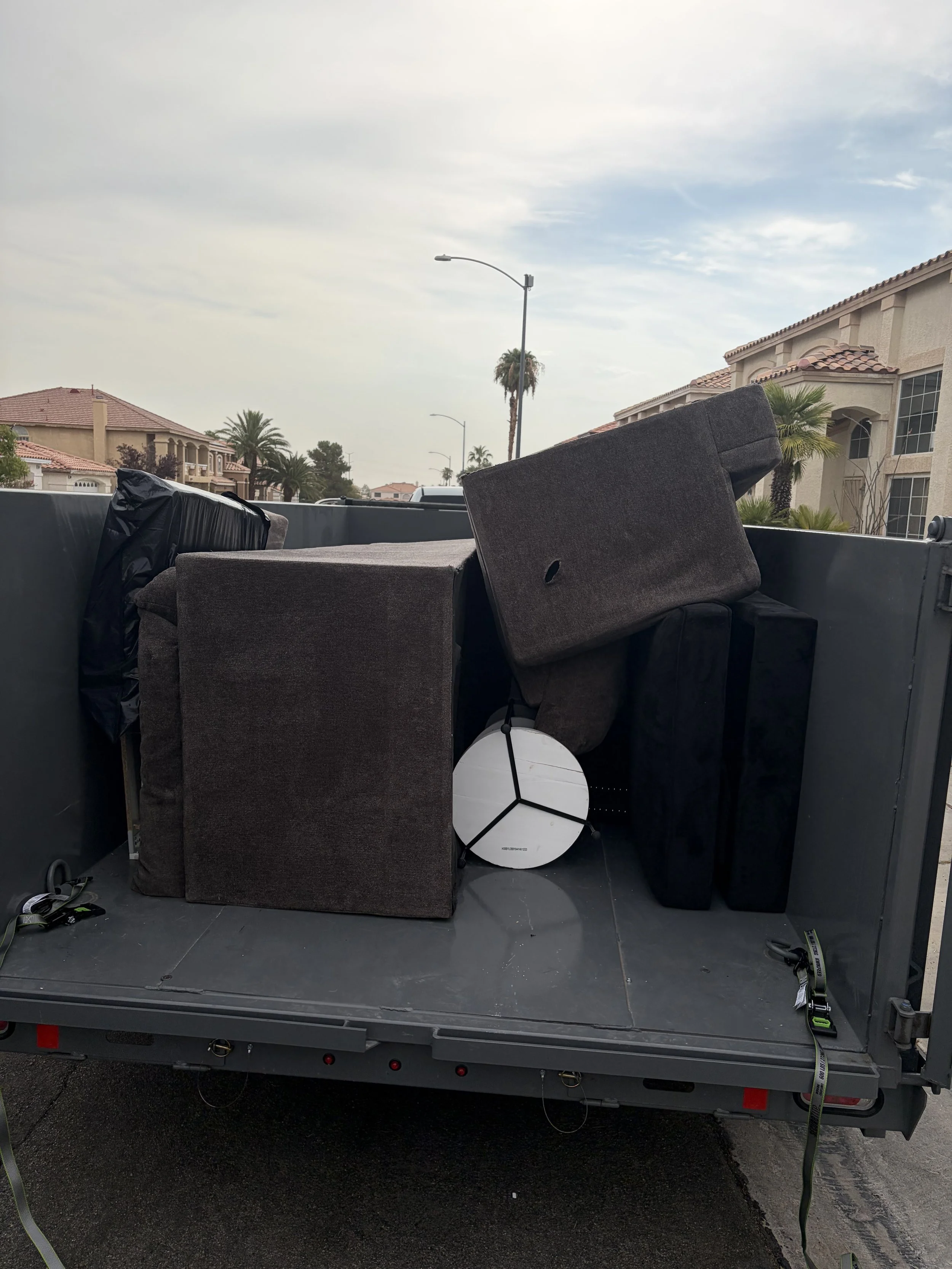 Back of a flatbed truck with dismantled furniture, a white round light fixture, and miscellaneous items including a black bag and tools, against residential houses and cloudy sky background.