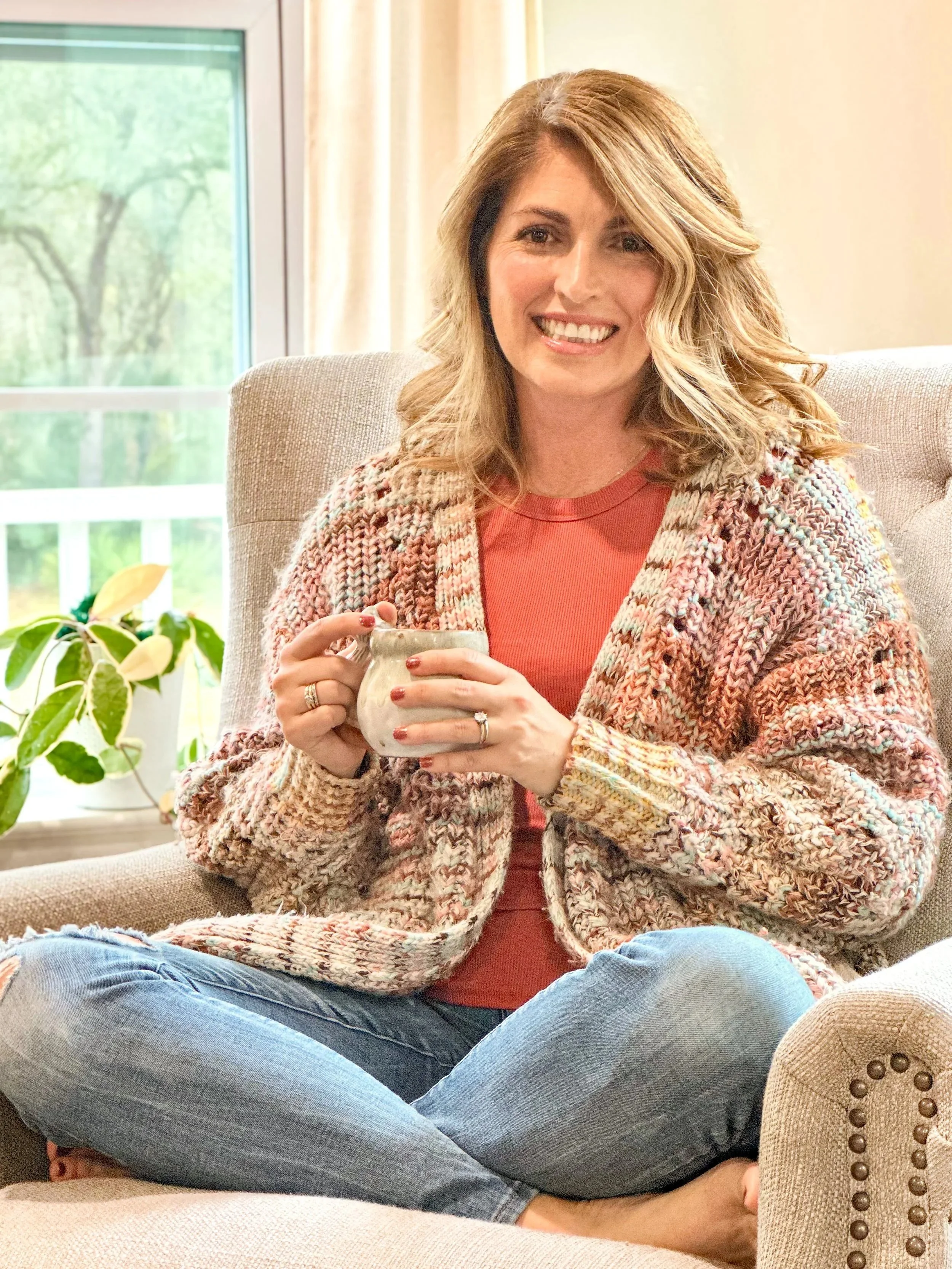 A woman with wavy blonde hair sitting on a beige couch, holding a cup, smiling, and wearing a patterned sweater and red shirt, in a room with a window and some plants.