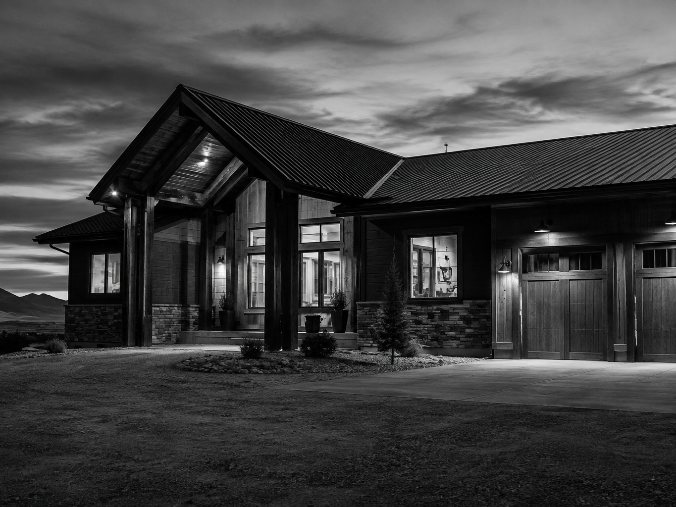 A black and white photo of a modern house with large windows and a metal roof, illuminated from inside, at dusk or night with cloudy sky and mountain landscape in the background.