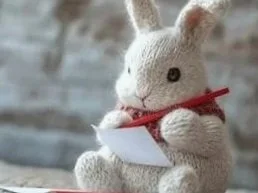 A white stuffed rabbit holding a piece of paper with a red ribbon around its neck.