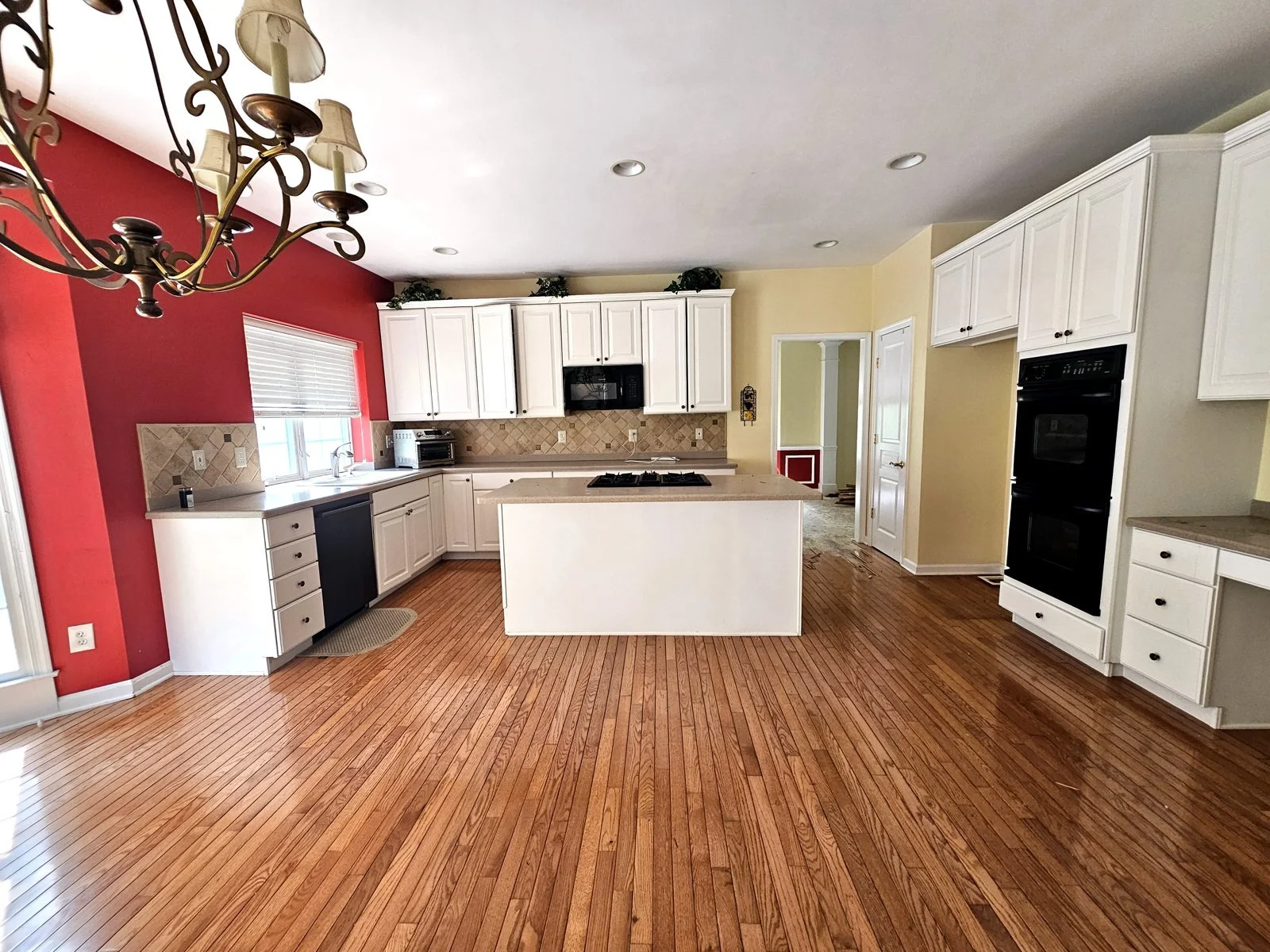 A spacious kitchen with white cabinets, a central island, a black built-in oven, and a red accent wall. Hardwood flooring and a chandelier are visible.