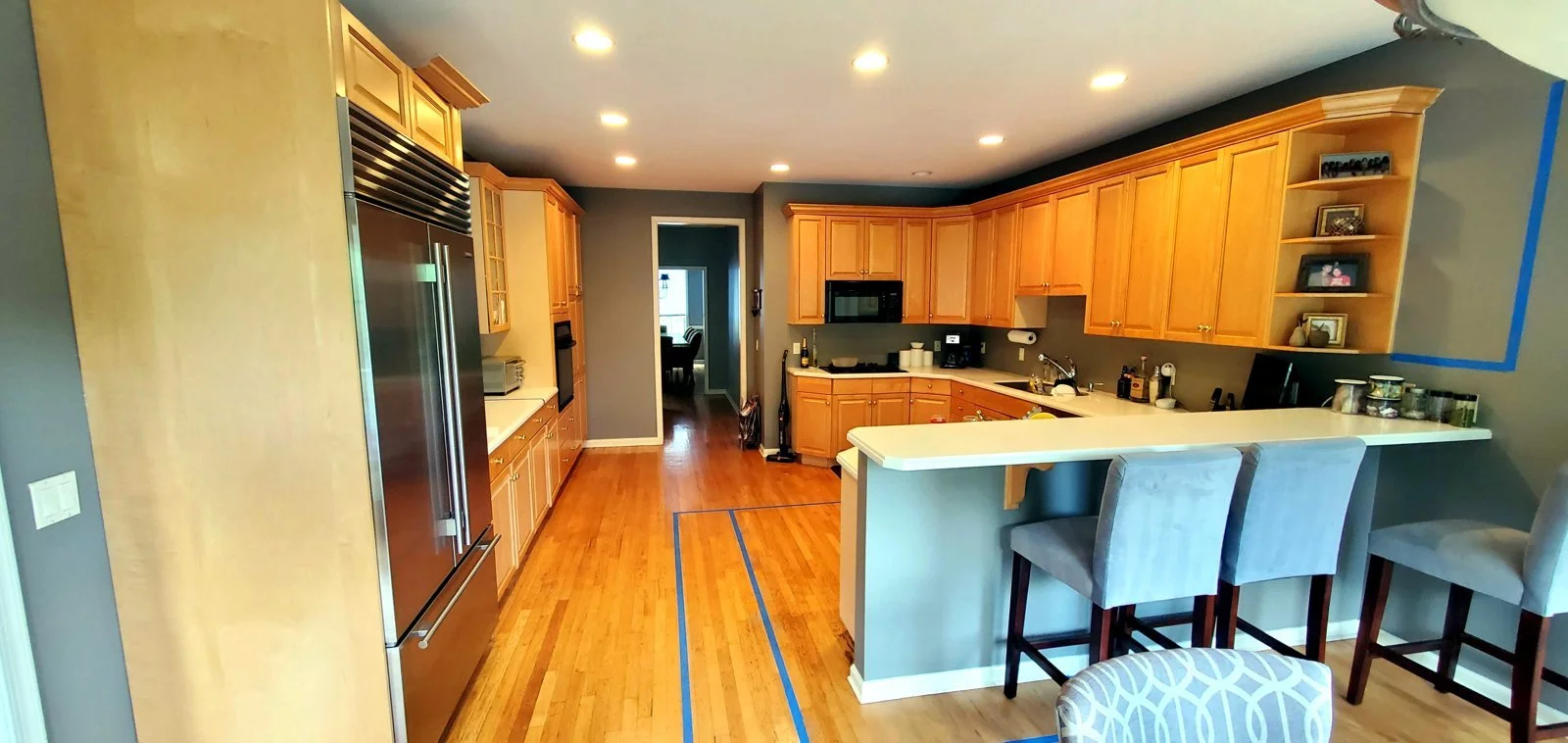 Modern kitchen with wooden cabinets, stainless steel refrigerator, gray walls, hardwood floor, and breakfast bar with gray upholstered chairs.