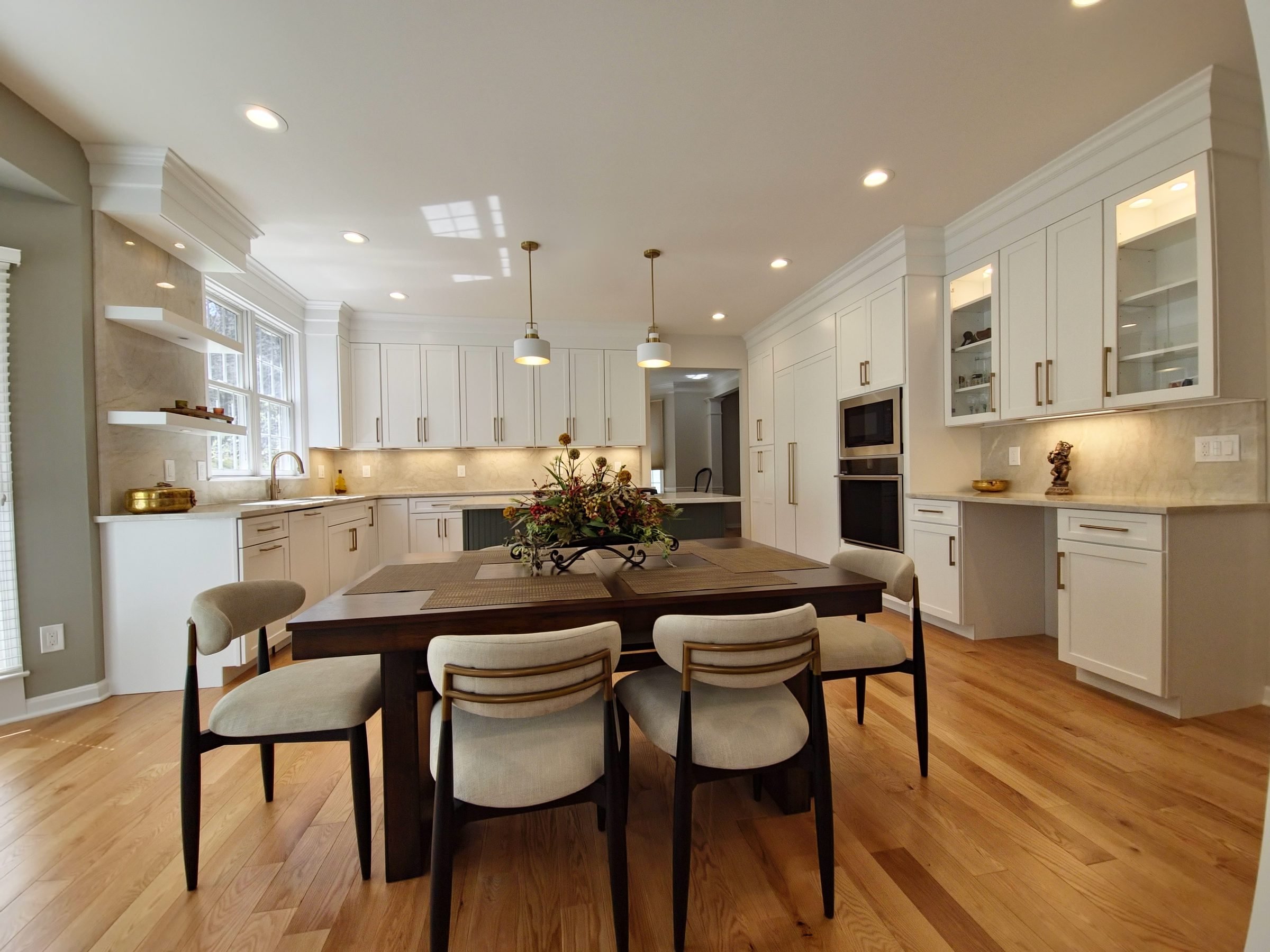 A spacious kitchen with white cabinets, a central island, and a dark wood dining table surrounded by beige upholstered chairs. The kitchen has large windows, hardwood floors, and modern white pendant lights.