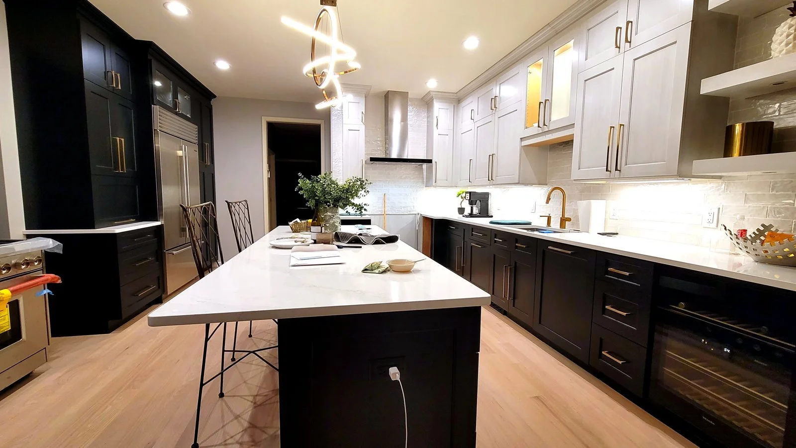 Modern kitchen with black lower and white upper cabinets, a white marble island with place settings and a green plant, hardwood floors, and a stainless steel refrigerator.