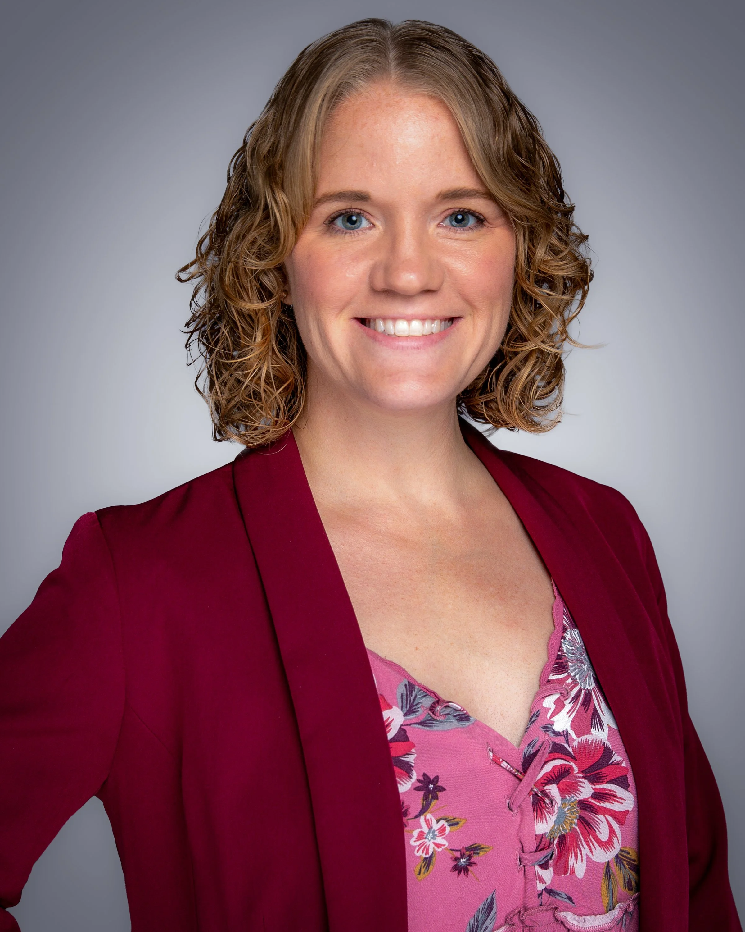 Close-up of a woman with curly blonde hair, smiling, wearing a dark pink blazer over a floral blouse.