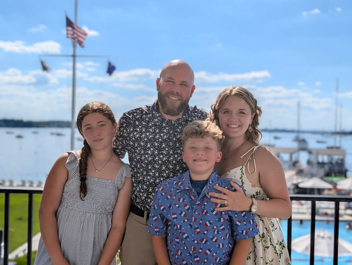 A family of five standing outdoors on a balcony near a marina with boats, American flags, and a bright blue sky in the background. They are smiling and dressed in casual summer clothing.