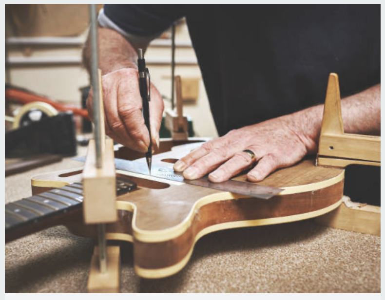 A person is working on building or repairing an acoustic guitar, measuring and marking the guitar's body on a workbench in a workshop.
