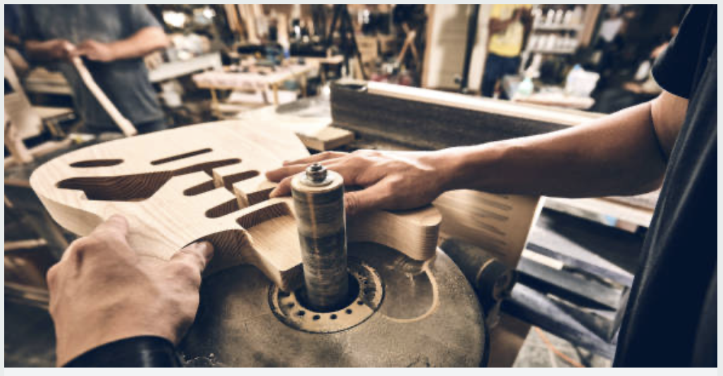 A person is working on shaping an acoustic guitar body in a woodworking shop, using a rotary sander to smoothen the wood.