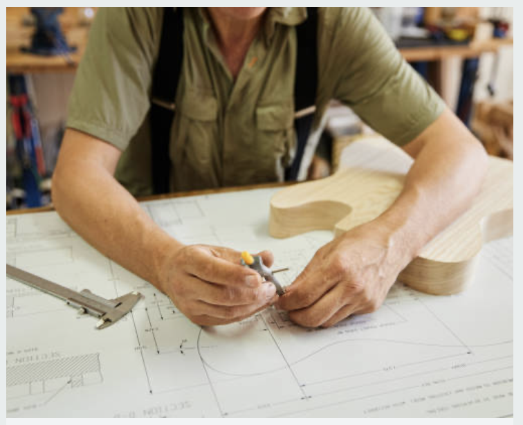 Person working on a woodworking project, drawing measurements on a blueprint with a pencil, with a wooden guitar body on the table.