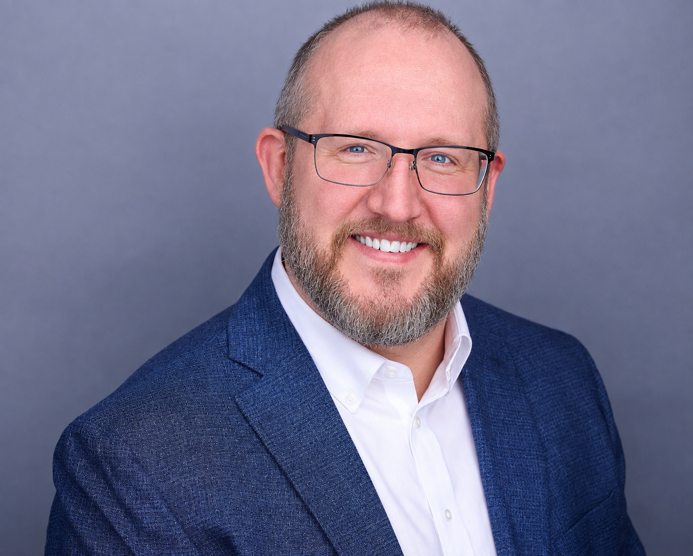 A man with glasses, a beard, and a smile, dressed in a white shirt and navy blazer, posing against a plain gray background.