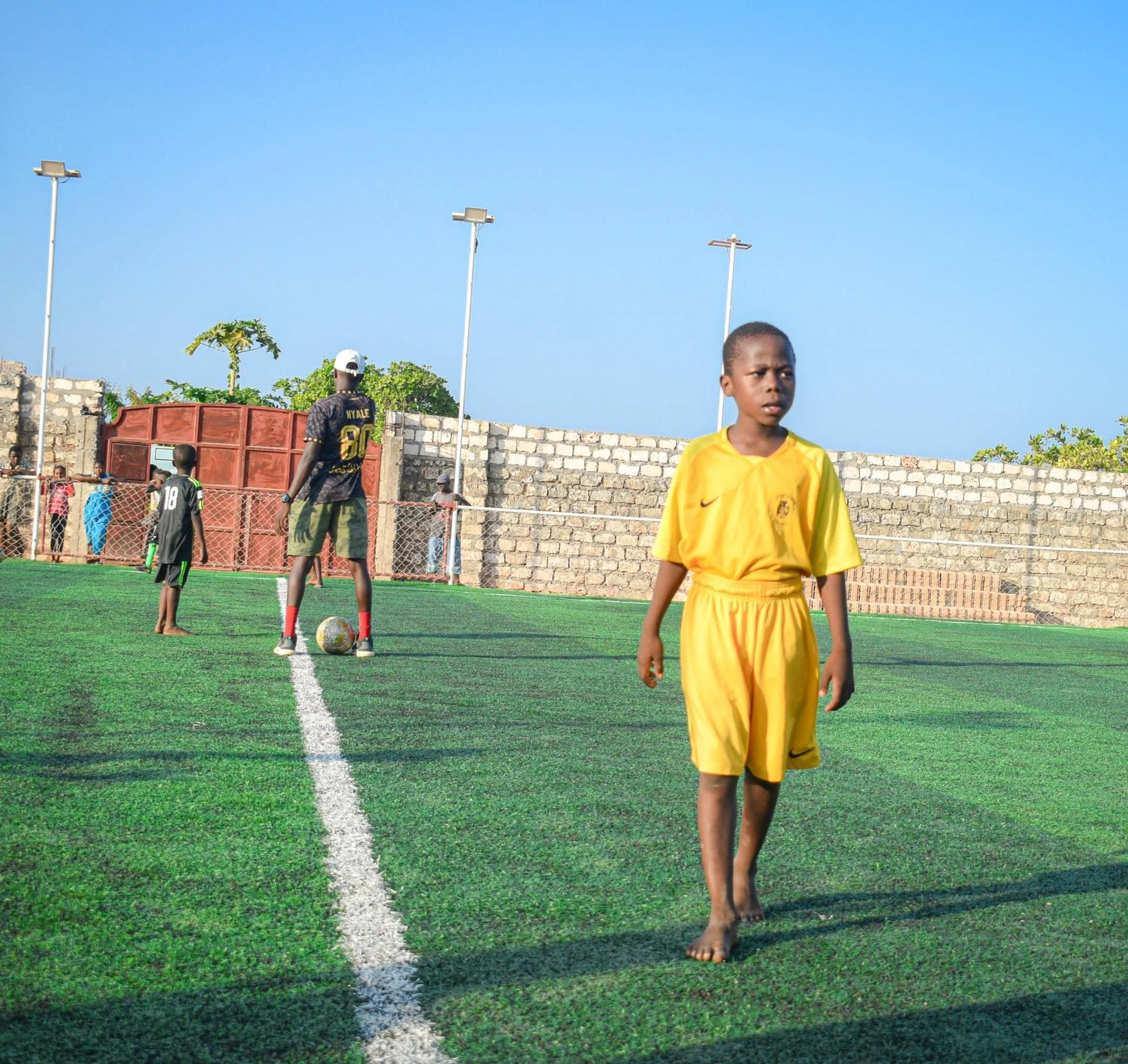 Young boy in yellow soccer uniform walking barefoot on synthetic soccer field. Background shows a brick wall, trees, and a blue sky with streetlights and several children and adults near a red gate.