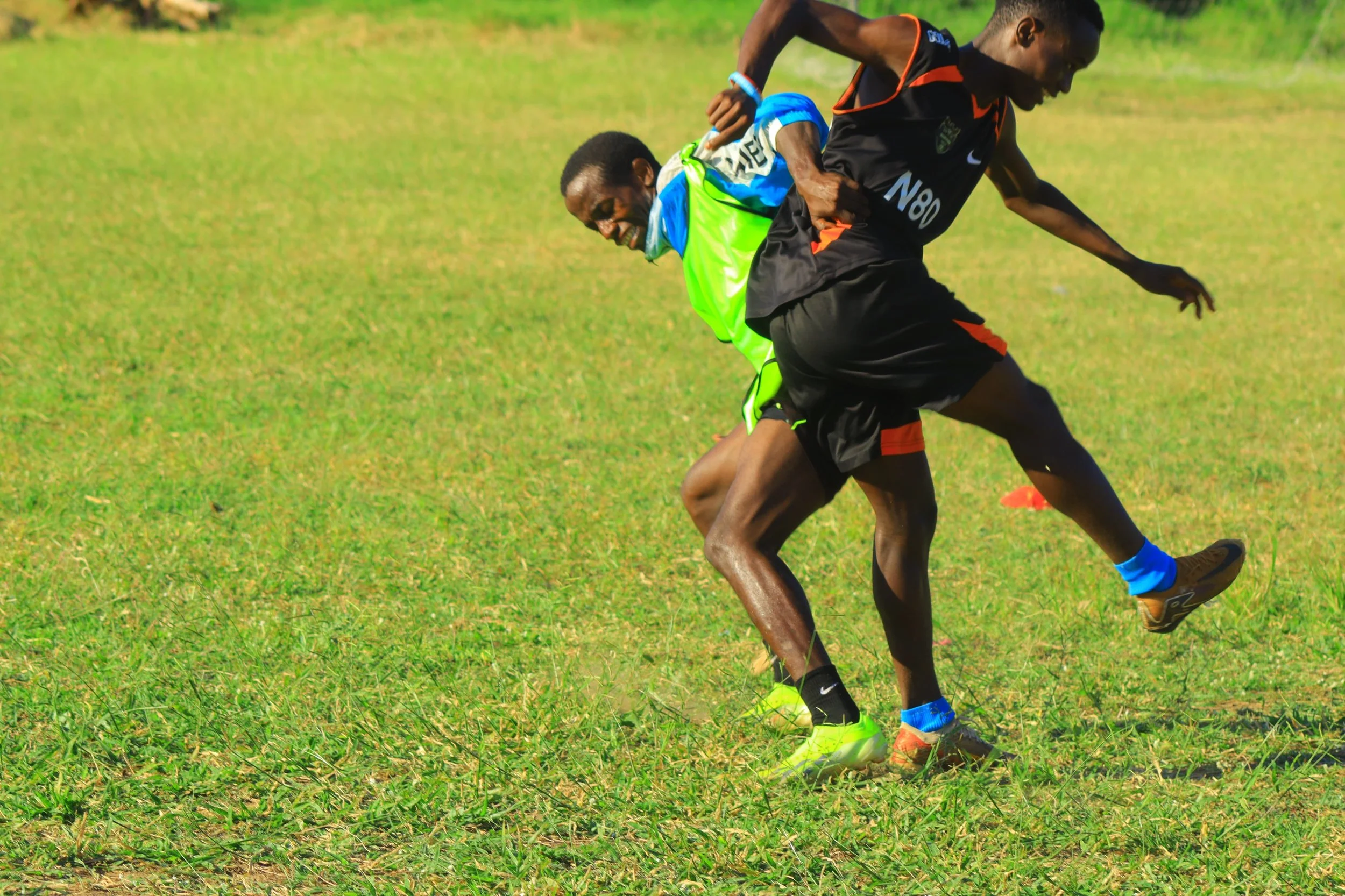 Two male runners racing on a grassy field, mid-stride, with expressions of determination, wearing athletic clothing and shoes, in Kilifi, Kenya. Wearing cleats. Football and growth and determination.
