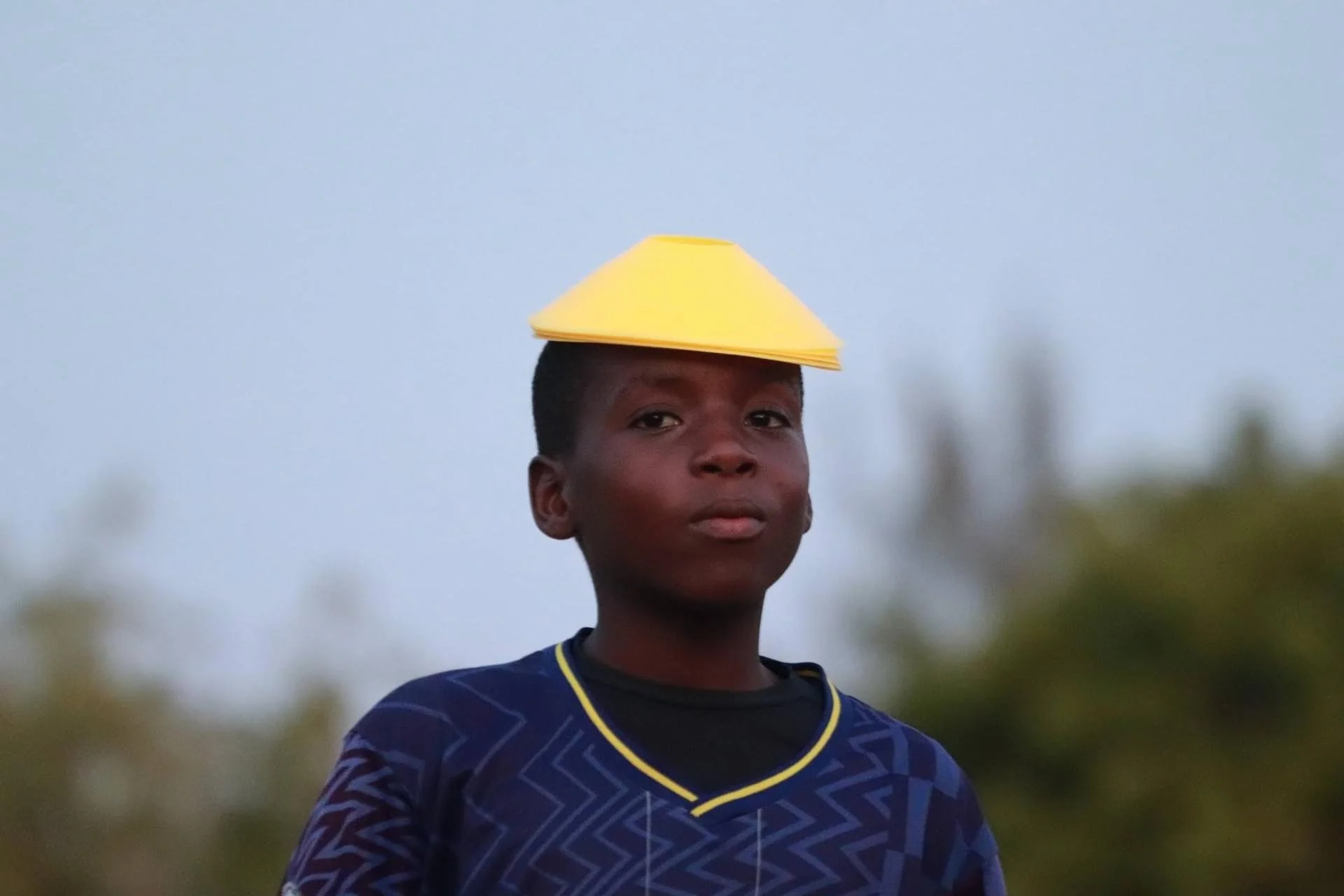 A young boy wearing a dark blue sports jersey with yellow trim and a yellow paper hat on his head outdoors against a blurred background of trees and sky.
