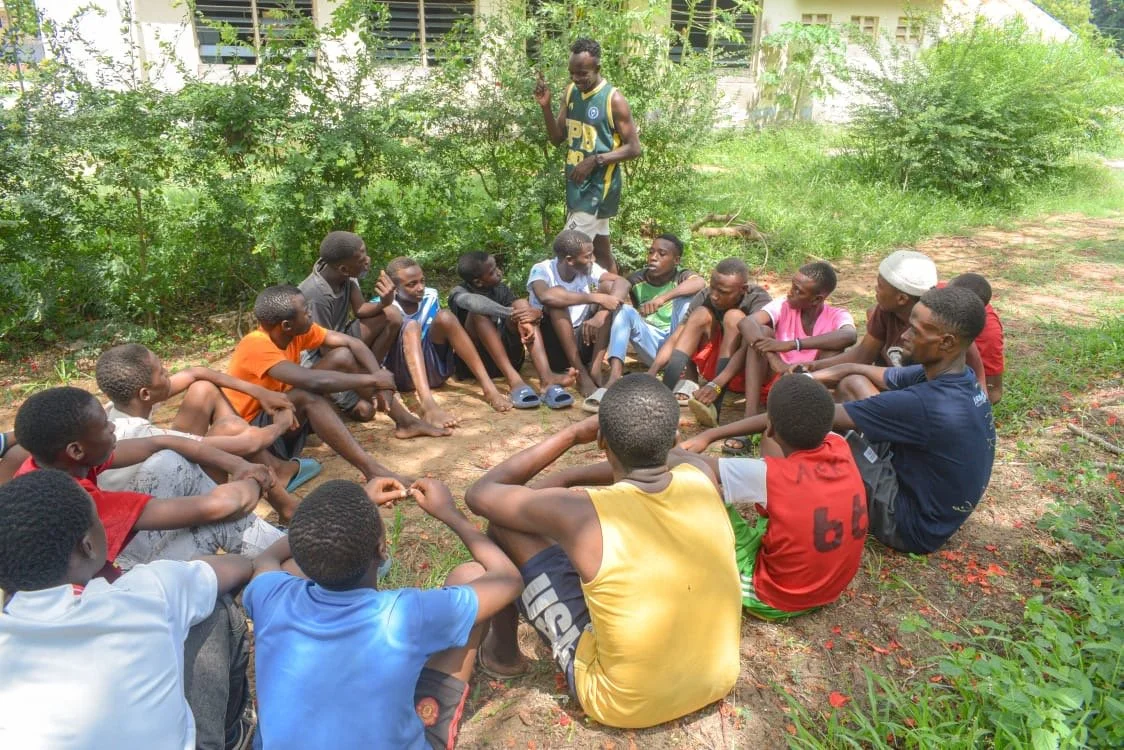 A group of children sitting in a circle outdoors, with one boy standing in the middle, surrounded by green foliage and trees. Group learning and development of skills by professional Nyale. 