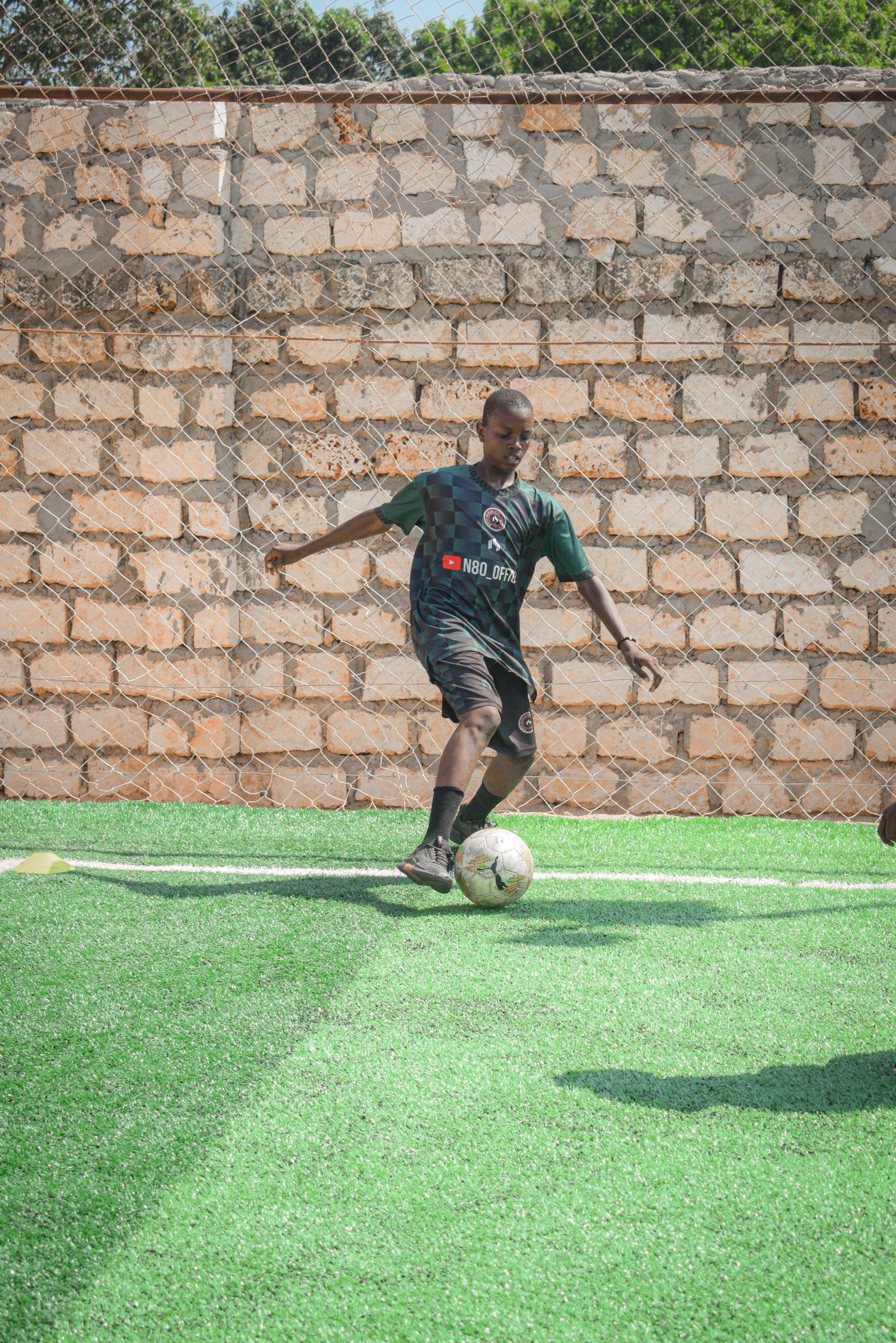 A young boy in a green and black sports jersey and shorts is playing soccer on a green artificial turf field, with a brick wall and chain-link fence in the background.