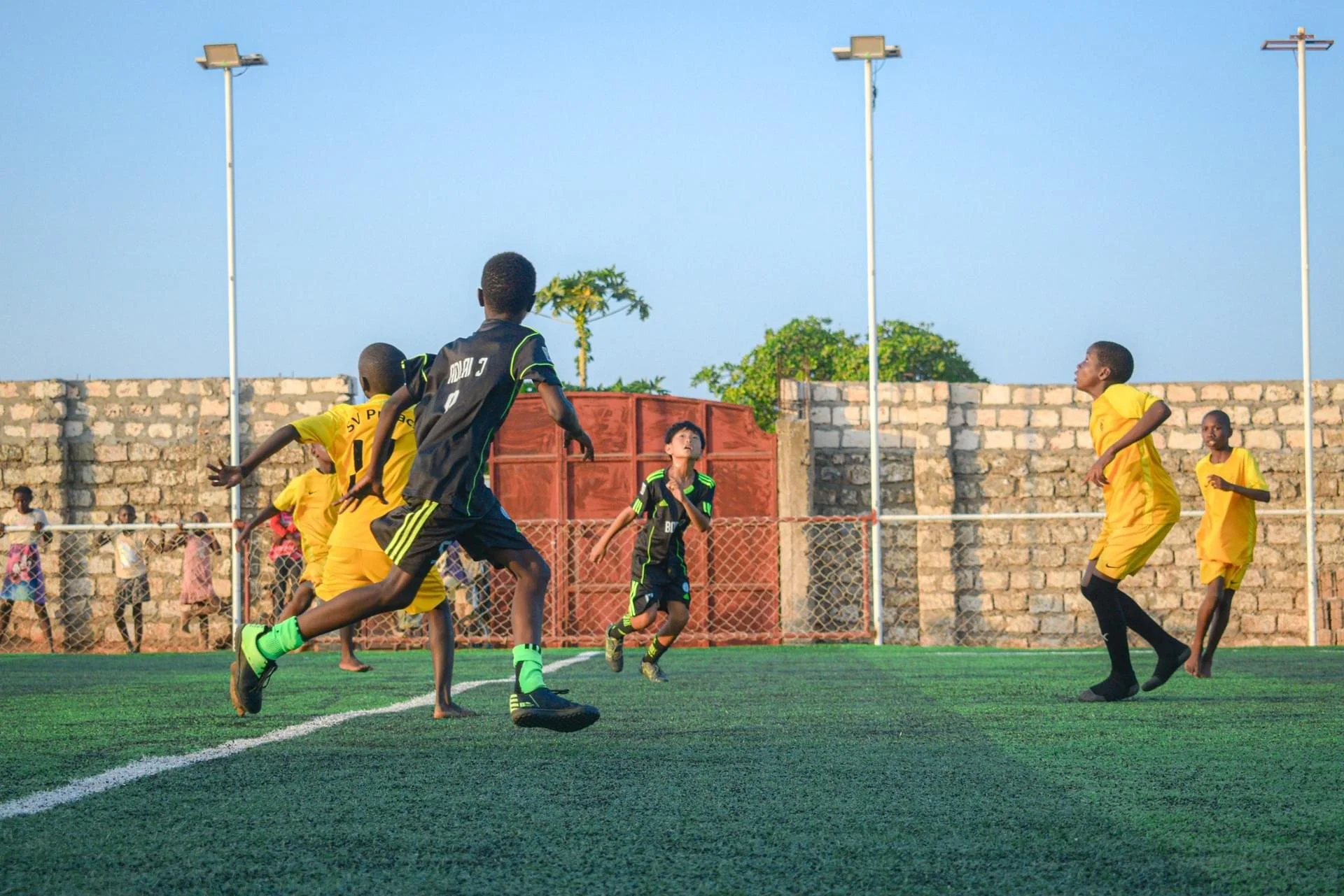 Children playing soccer on an outdoor field with a brick wall and trees in the background, under a clear blue sky.