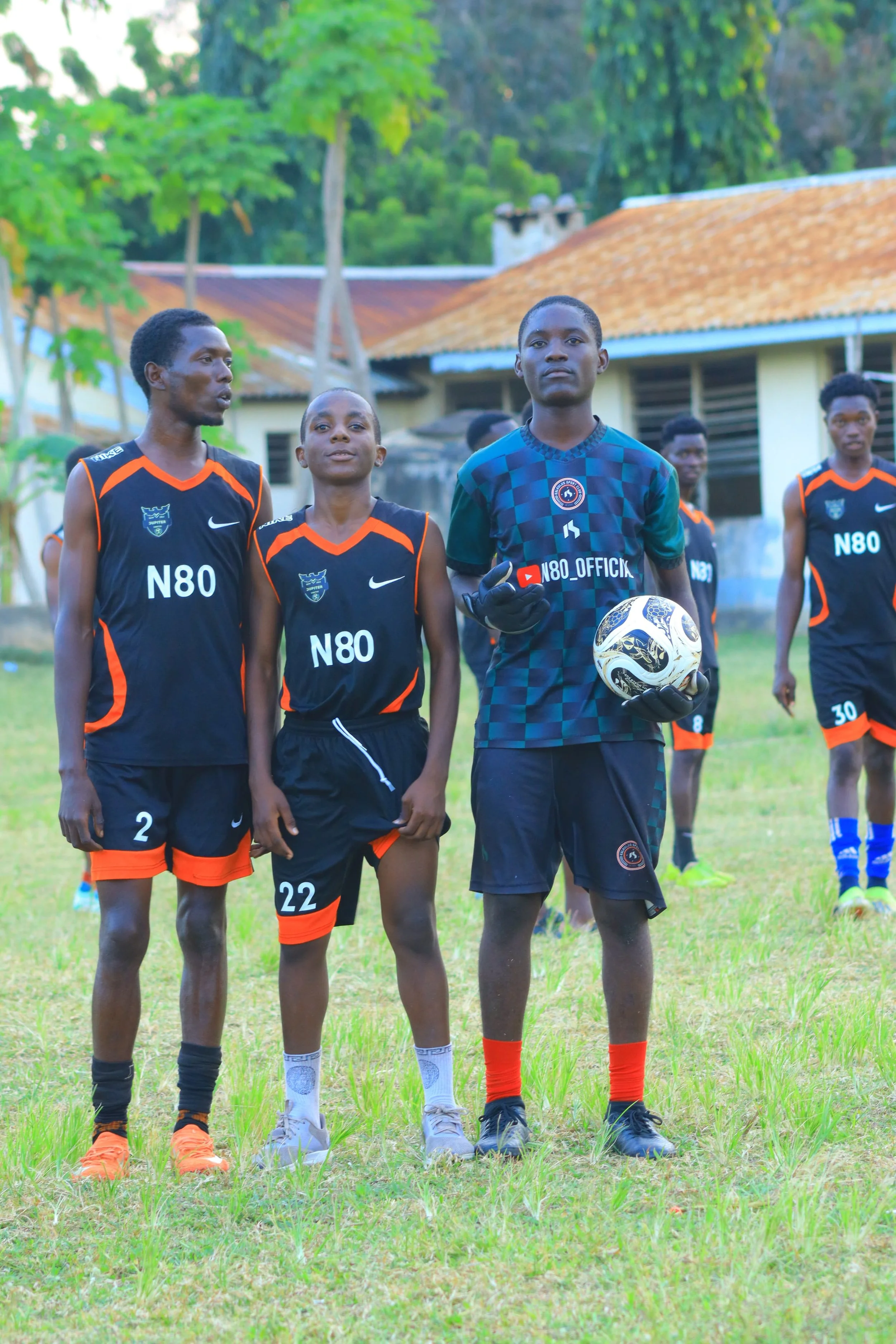 A group of young soccer players, wearing black and orange uniforms, standing on a grassy field. One player, wearing a goalkeeper jersey, is holding a soccer ball. In the background, there are trees and a building with an orange roof.