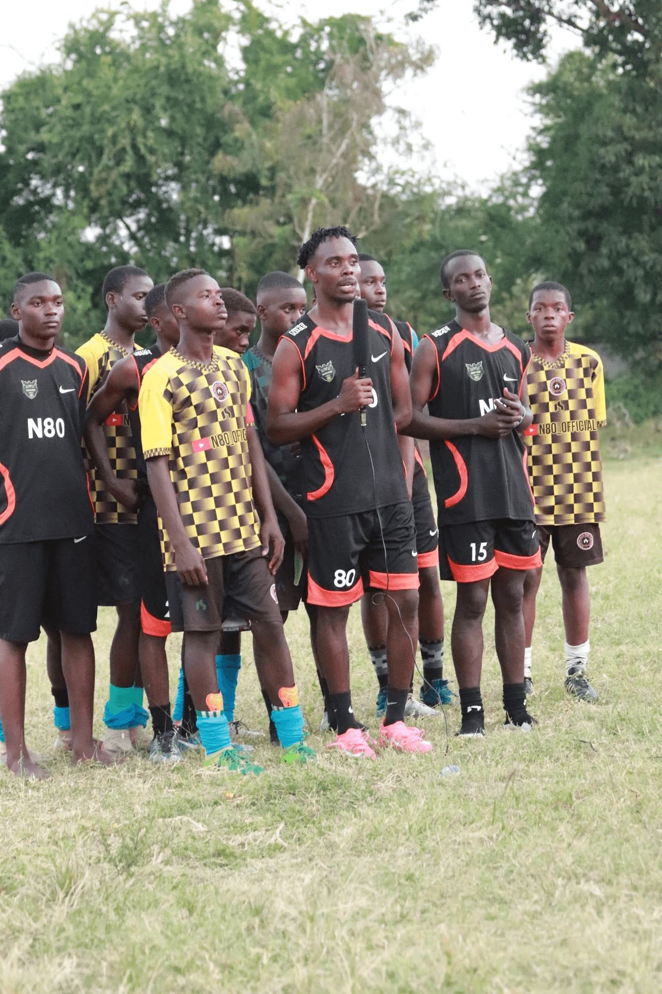 A group of young athletes standing outdoors on a grassy field, wearing sports uniforms in black and yellow, with one man in the center holding a microphone and speaking to the group.