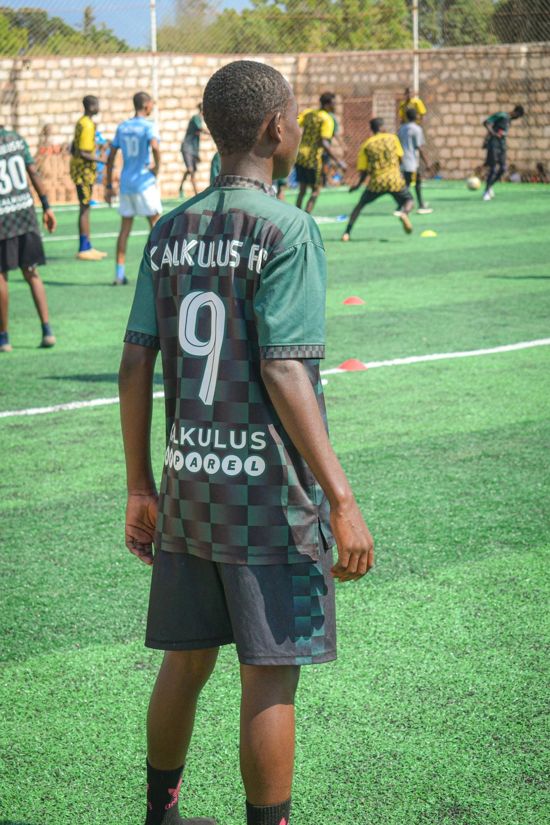 A young soccer player stands on a green field, wearing a green checkered soccer jersey with the number 9 on the back and the text 'CALKULUS FC.' The player is observing their teammates practicing in the background.