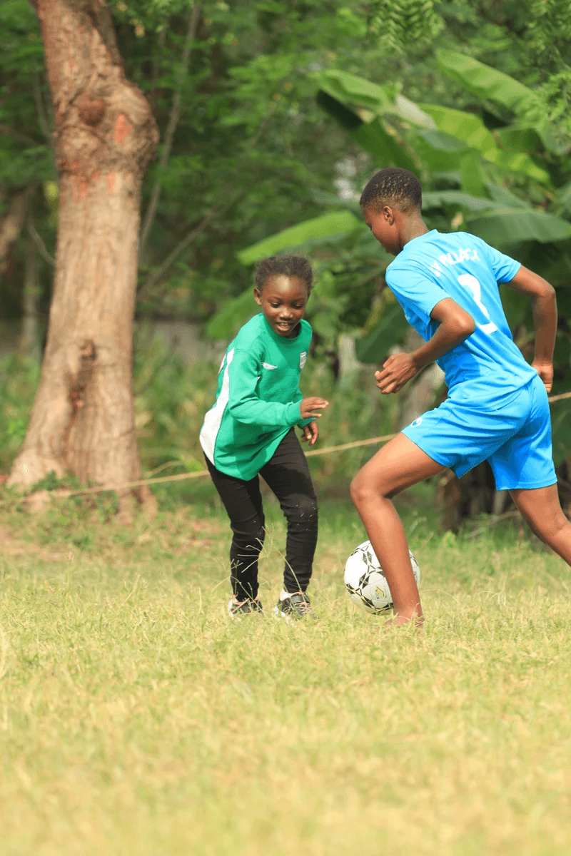 Two children playing soccer outdoors, one in a green jersey and the other in a blue jersey, with a soccer ball on the grass in a natural, green environment in Kilifi, Kenya.