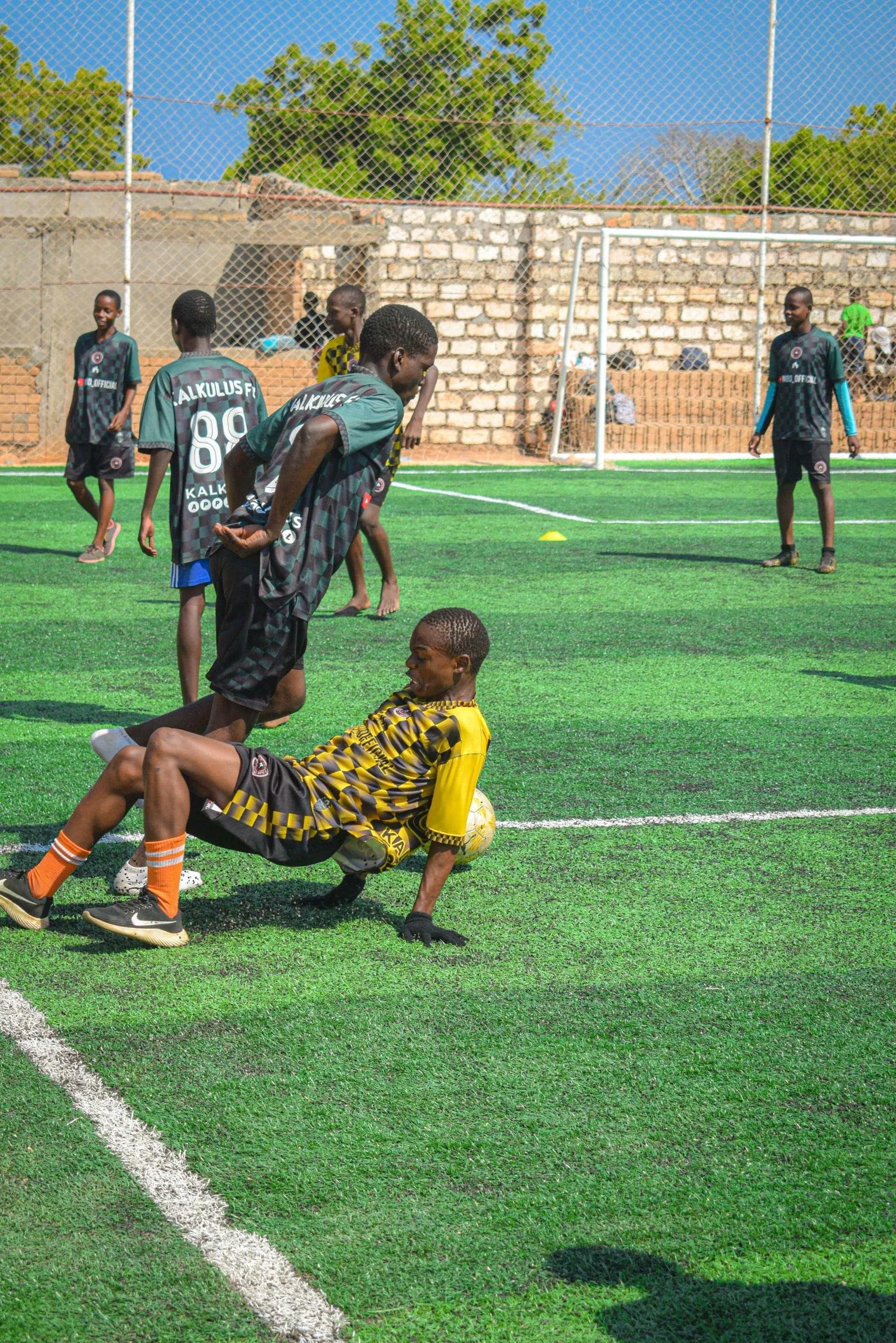 A group of young boys playing soccer on a small artificial turf field, with a brick wall and a chain-link fence in the background, during daytime.