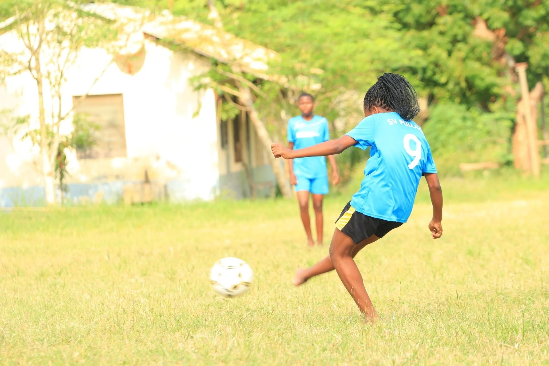 A young girl in a blue sports jersey kicking a soccer ball on a grassy field, with a boy and greenery in the background.