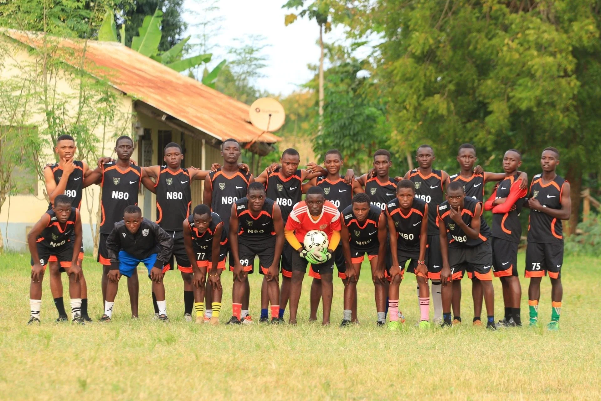 Group of young boys in black and red soccer uniforms posing for a team photo on a grassy field, with some holding a soccer ball and others standing with arms around each other, in front of trees and a yellow building in Kilifi, Kenya.