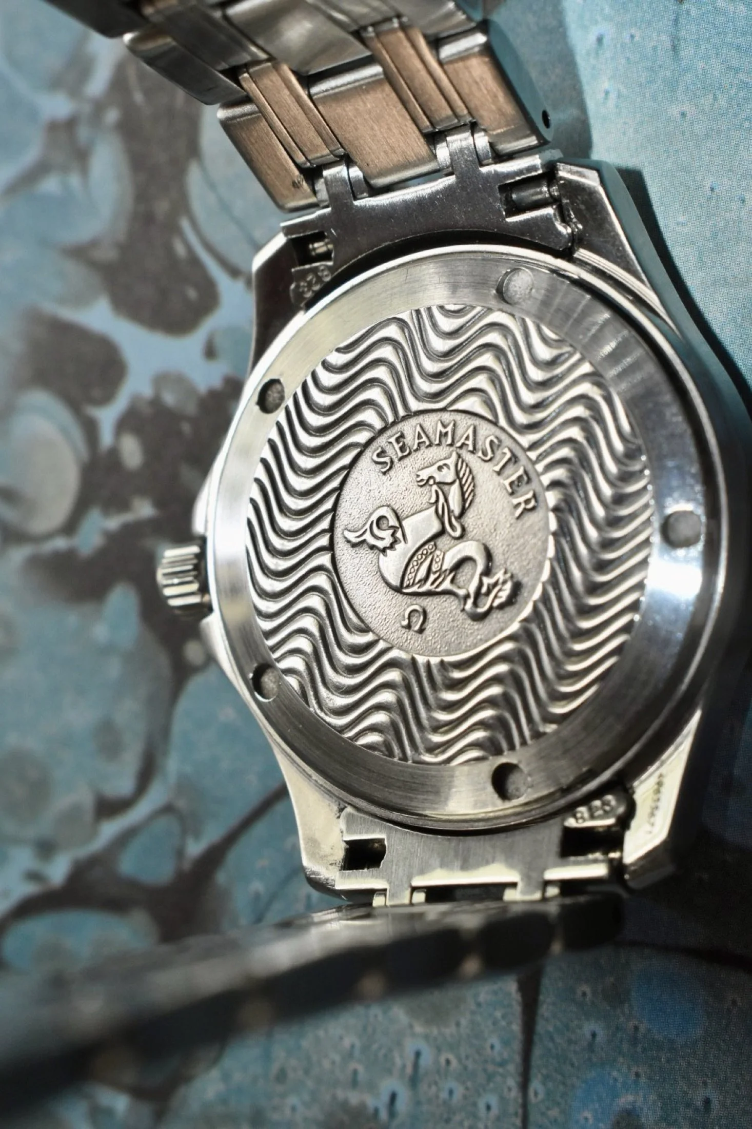 Close-up of the back of a stainless steel Omega Seamaster wristwatch, showing the engraved logo and patterned design on the case back.
