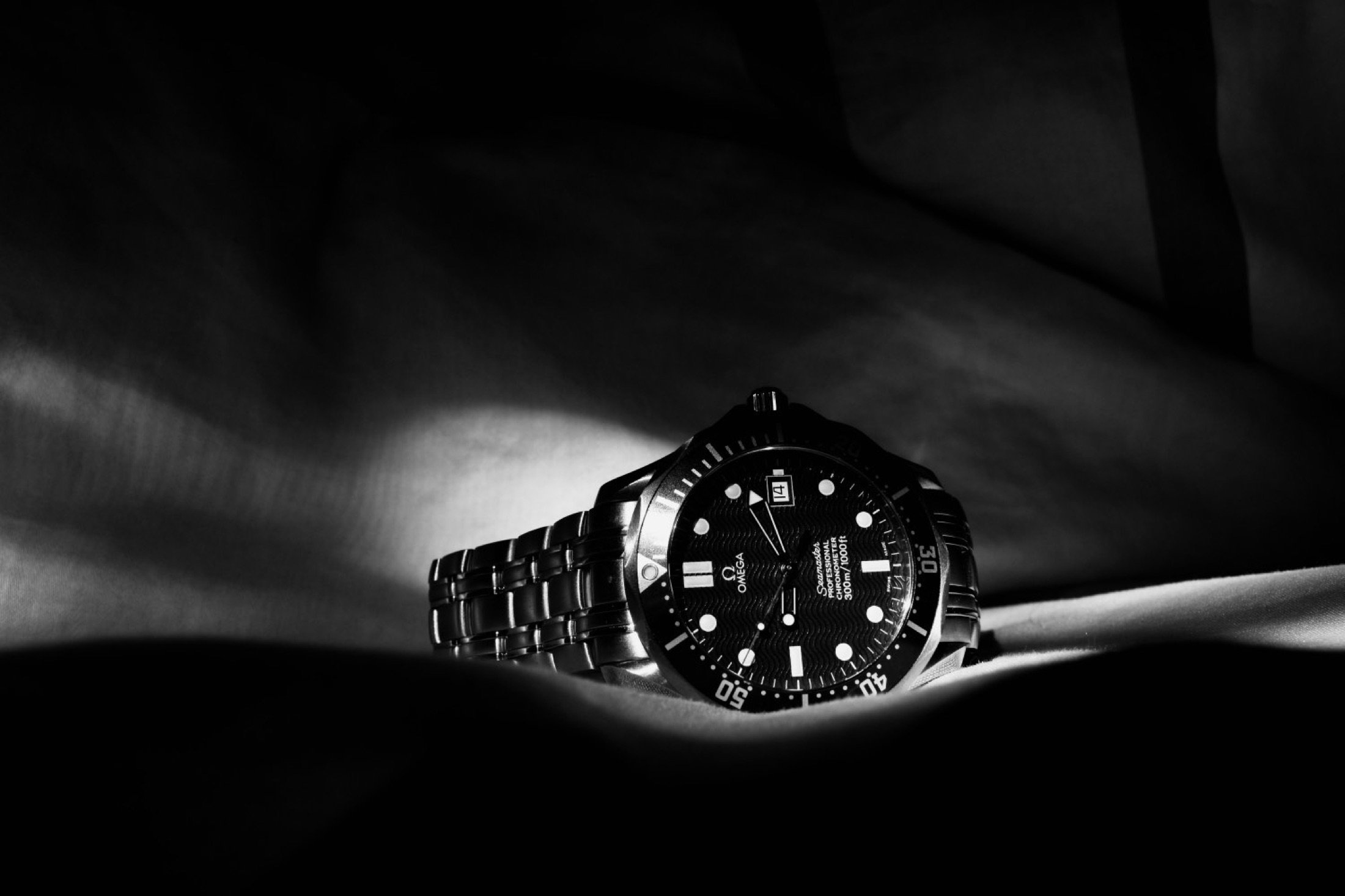 A close-up of a black stainless steel Omega Seamaster professional dive watch with a black textured dial and bezel, illuminated by a beam of light, against a dark background.