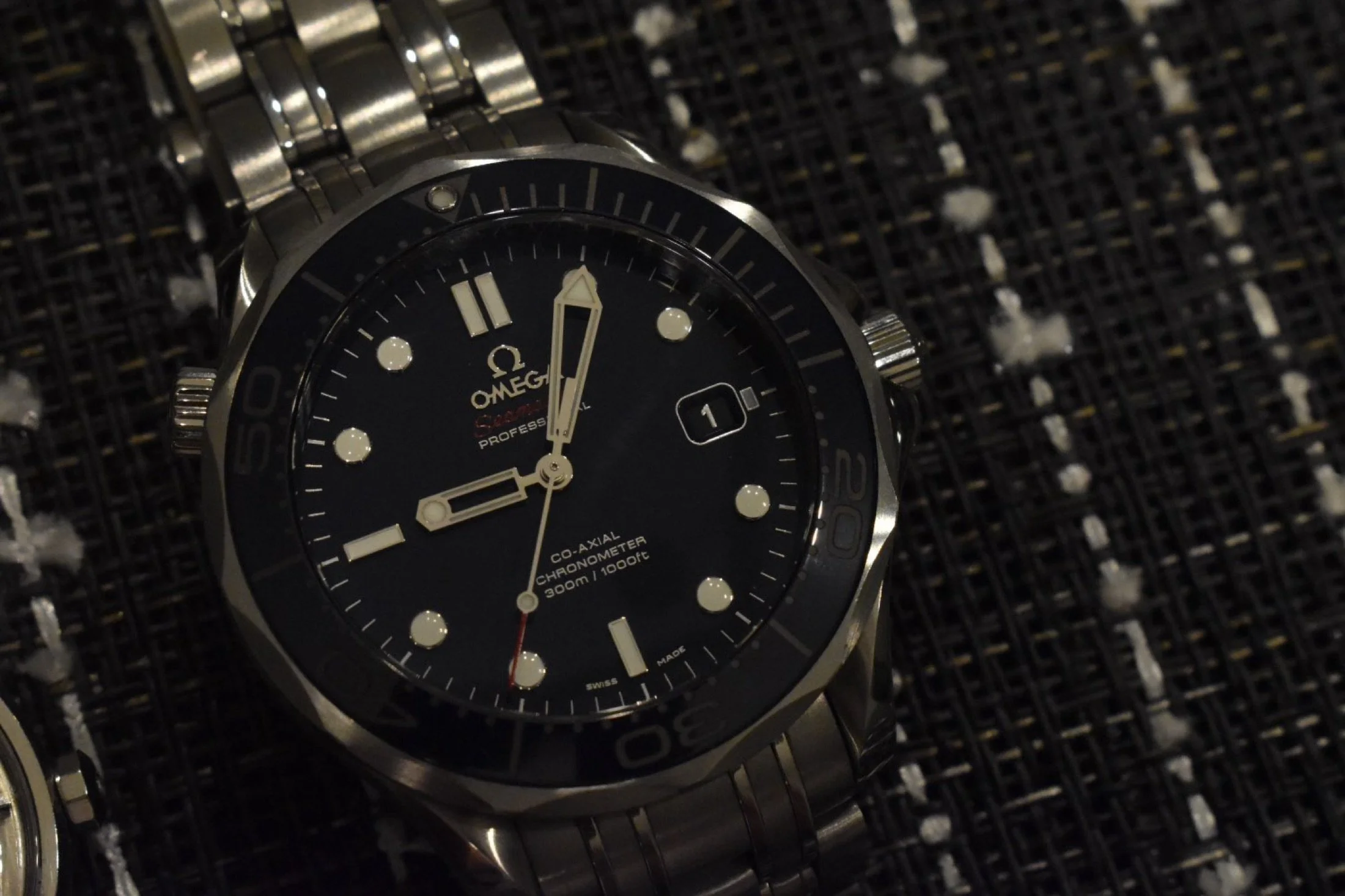 A close-up photo of an Omega wristwatch with a black dial and a metal band, placed on a textured black surface.