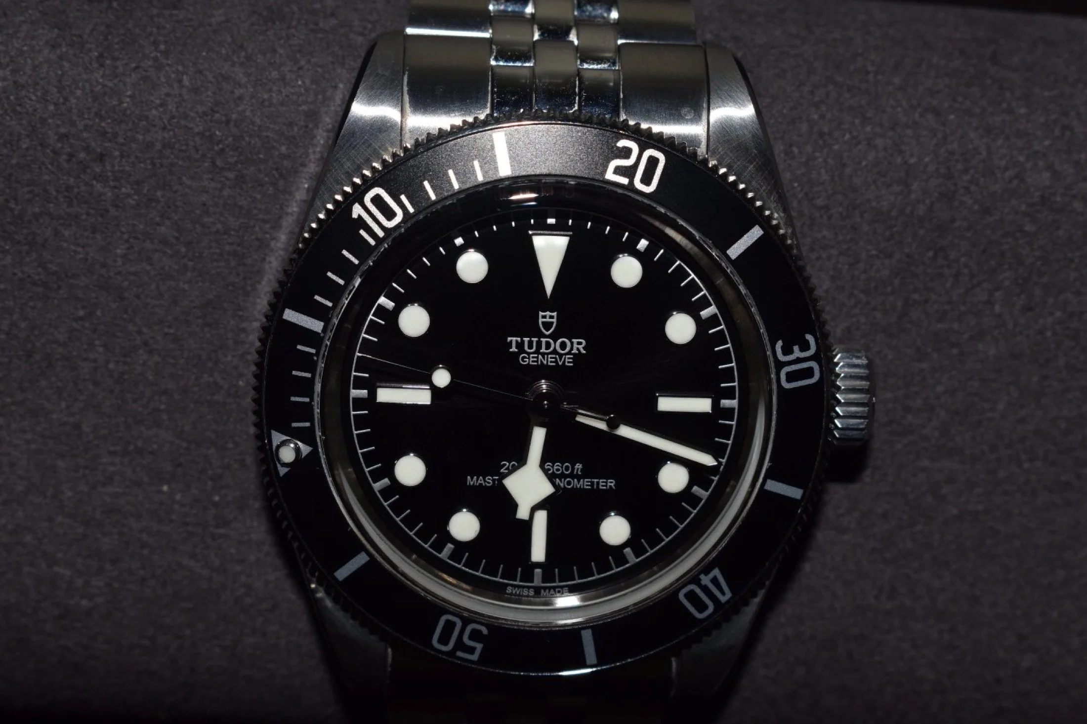 A close-up of a Tudor dive watch with a black dial, white markers, and a stainless steel bracelet, set against a dark background.