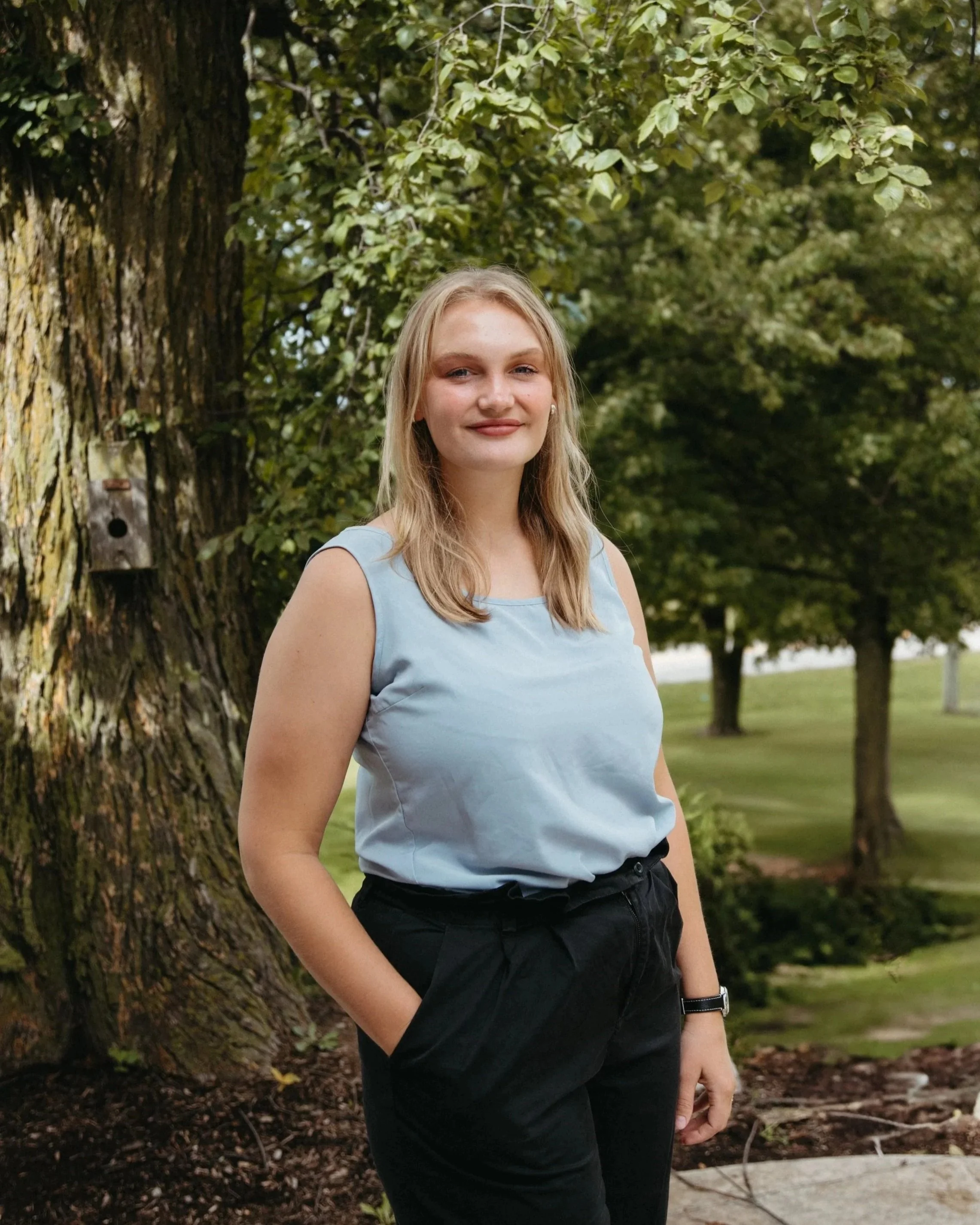 A young woman with blonde hair, wearing a light blue sleeveless top and black pants, is standing outdoors near a tree with green leaves. She is smiling and looking at the camera, with a park and trees in the background.
