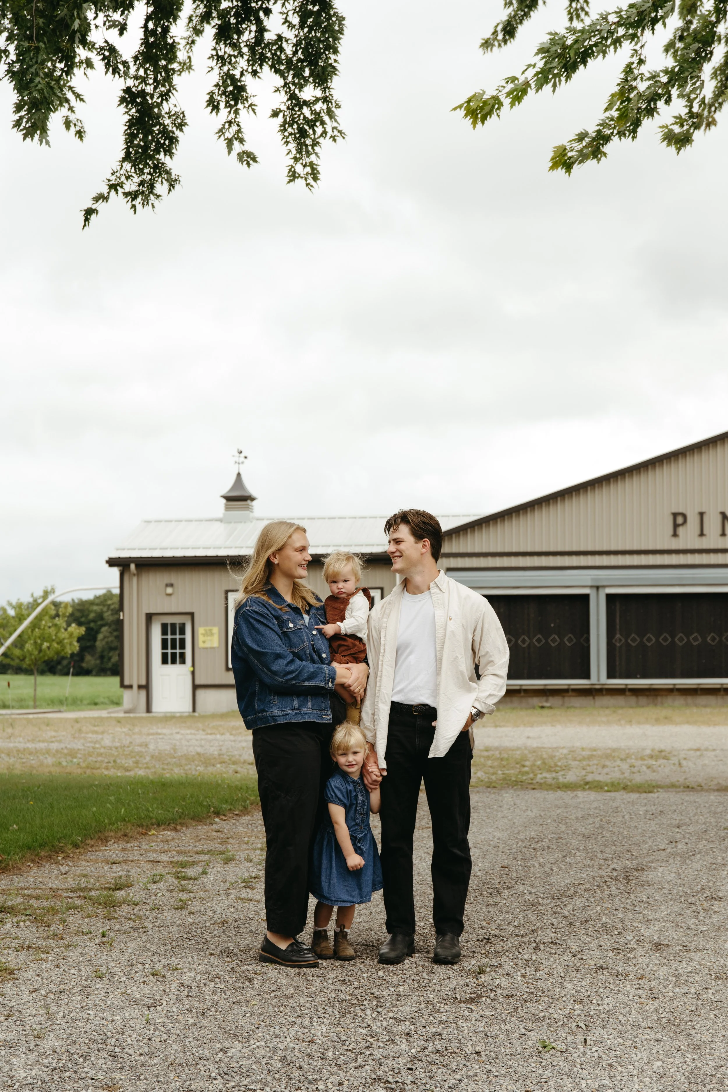 A family of five standing outdoors in front of a barn with a weather vane, on a cloudy day. The family includes a woman, a man, and three young girls, some of whom are holding hands and smiling.